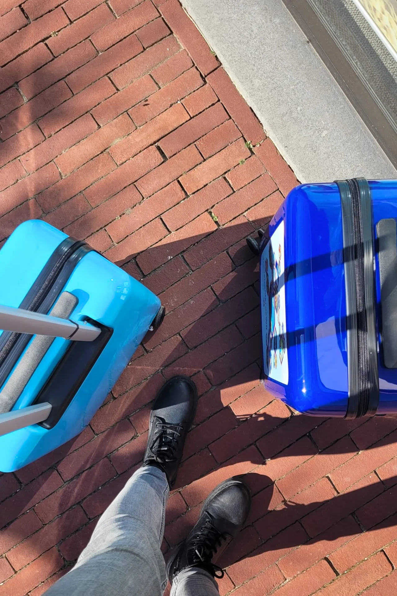 Bovenaanzicht van twee blauwe handbagage trolleys van Travelcenter de Wit, met een strak, modern ontwerp. Naast de trolleys zijn een grijze spijkerbroek en zwarte laarsjes zichtbaar, wat de casual, stijlvolle uitstraling van de set benadrukt. De koffers zijn lichtgewicht met vier wielen voor gemakkelijke manoeuvreerbaarheid, ideaal voor handbaggage tijdens vliegreizen.