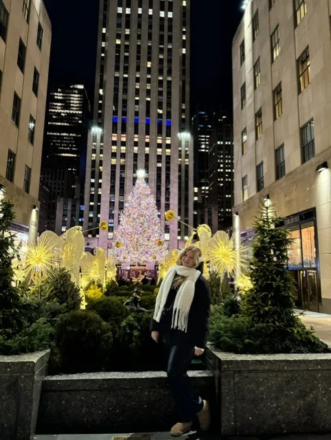 Een blonde vrouw poseert ’s avonds voor het Empire State Building in New York, met een prachtig verlichte kerstboom.