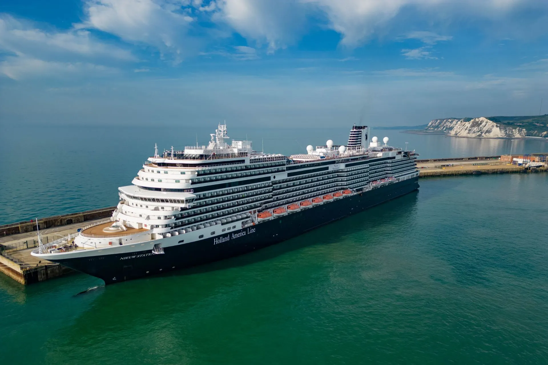 Cruiseschip Nieuw Statendam van Holland America Line aangemeerd in de haven van Dover, met uitzicht op de witte krijtrotsen en kalme zee.