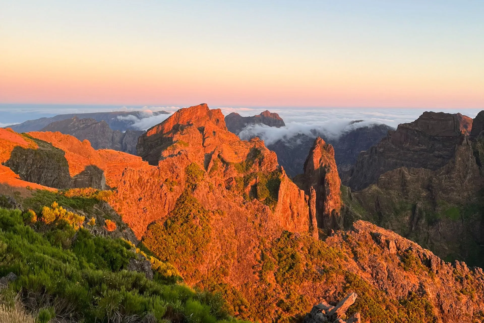 Fel verlichte bergtoppen bij Pico do Arieiro tijdens zonsopgang. De rotsen kleuren intens oranje door het eerste zonlicht, terwijl de lucht zachtblauw is en de horizon een lichte roze gloed toont. Wolken vullen de valleien tussen de bergen.