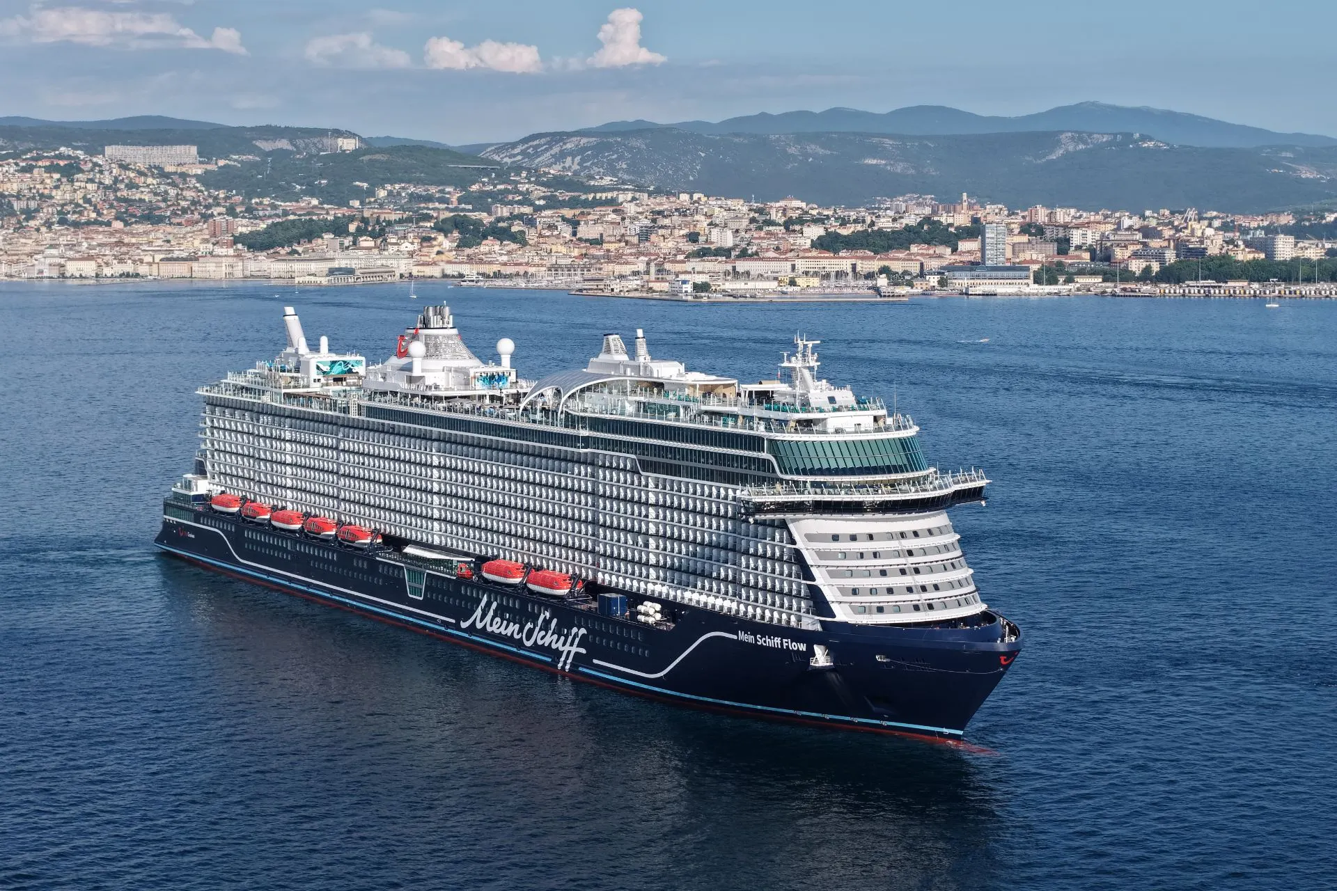 De Mein Schiff Flow vaart langs de kust van Triëst en laat goed de indrukwekkende omvang van het moderne cruiseschip zien. Met maar liefst 19 dekken, talloze balkonhutten en uitgebreide faciliteiten aan boord vormt dit nieuwste schip van Mein Schiff een comfortabele uitvalsbasis voor een cruise door de Middellandse Zee.