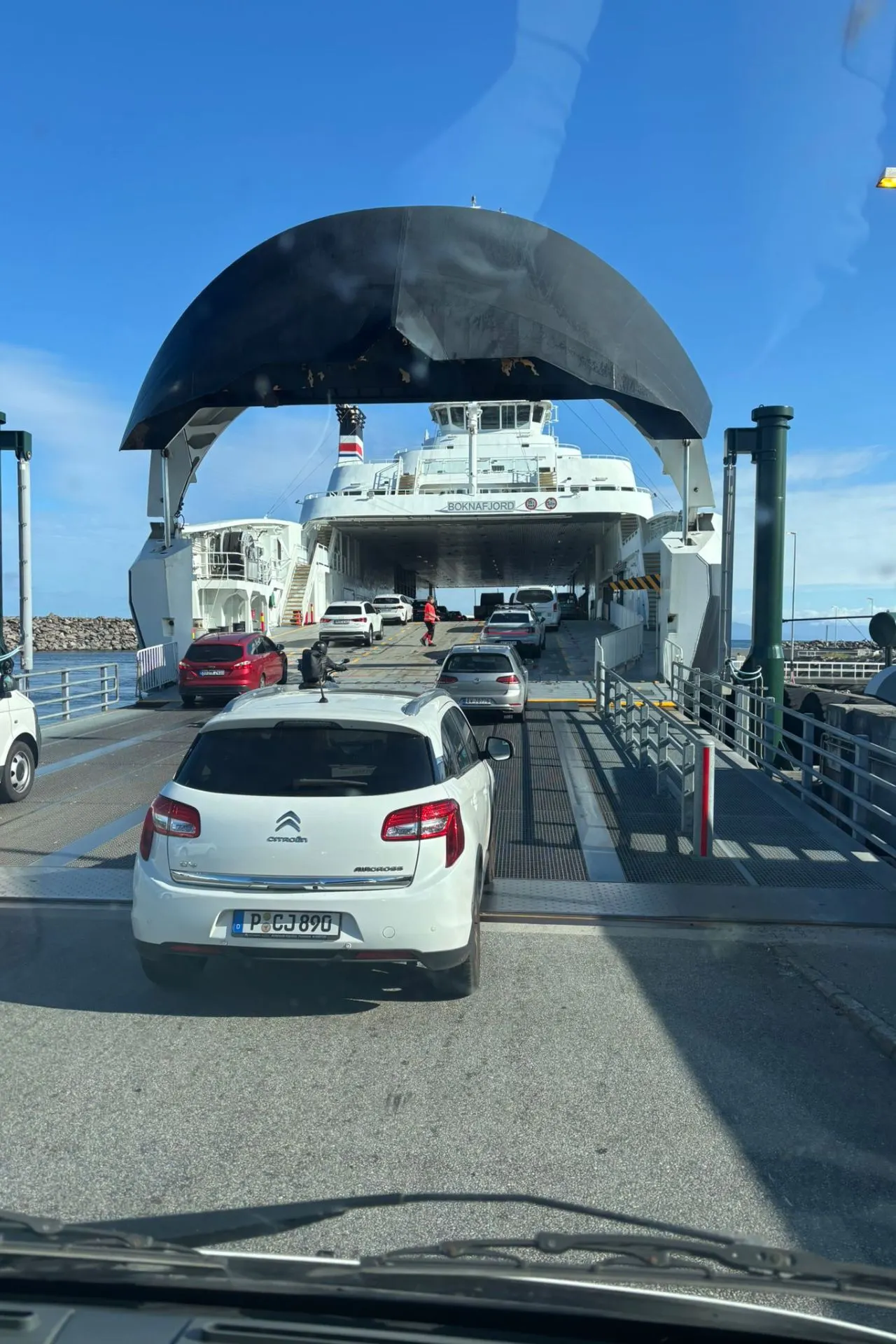 Auto's staan in de rij om een veerboot op te rijden. Op de voorgrond een witte Citroën met Frans kenteken, daarachter een rode auto. De veerboot heeft een grote zwarte boogvormige constructie boven de laadklep. Blauwe lucht en water op de achtergrond.