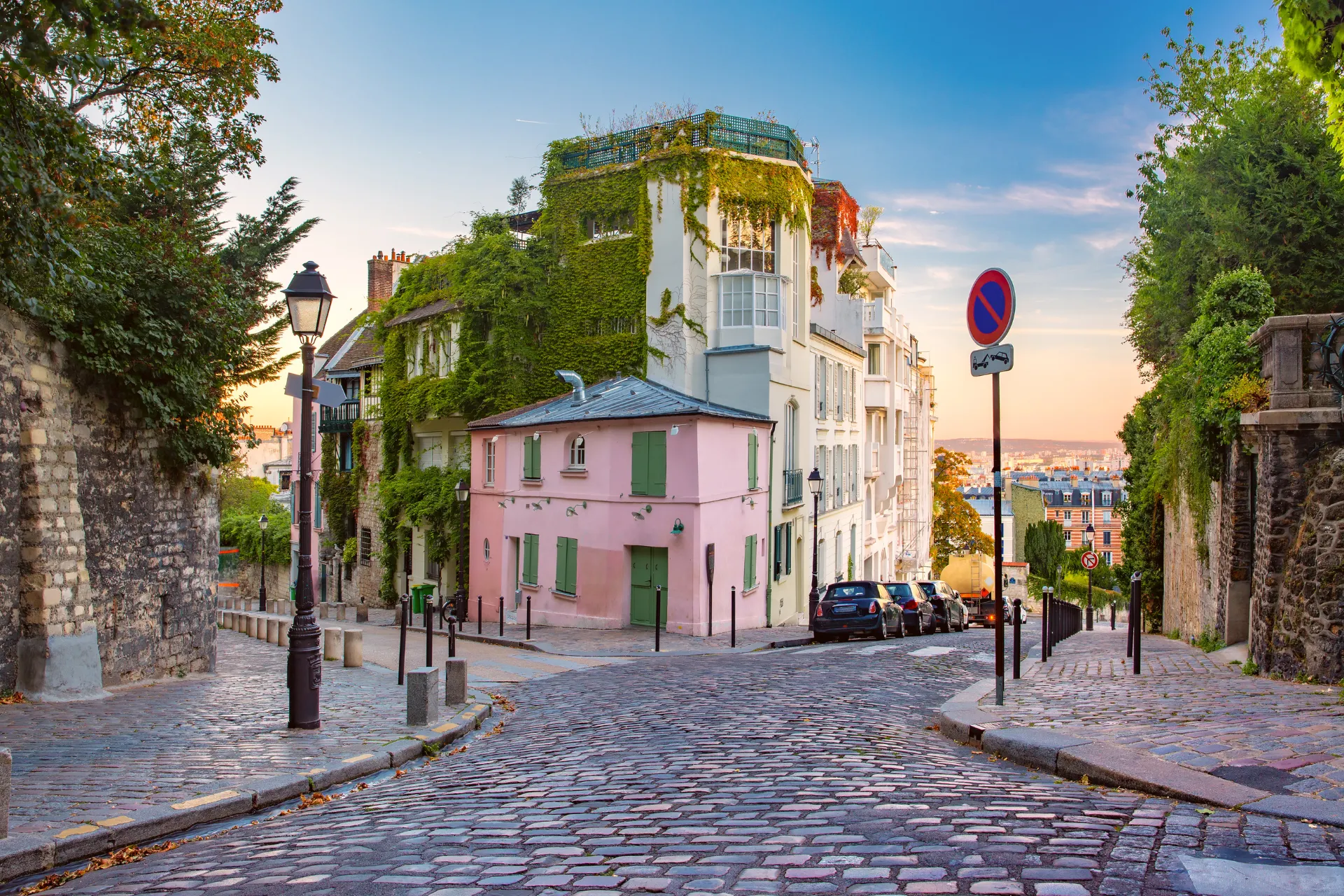 Charmante straat in Montmartre, Parijs, met geplaveide wegen en een pittoresk roze huis omgeven door groene klimop. Op de achtergrond zijn klassieke Parijse gebouwen zichtbaar, terwijl de zon een warme gloed over de scène werpt. Straatlantaarns en geparkeerde auto’s voegen een authentieke sfeer toe aan deze iconische wijk, populair bij toeristen voor kunst, cultuur en romantische wandelingen.