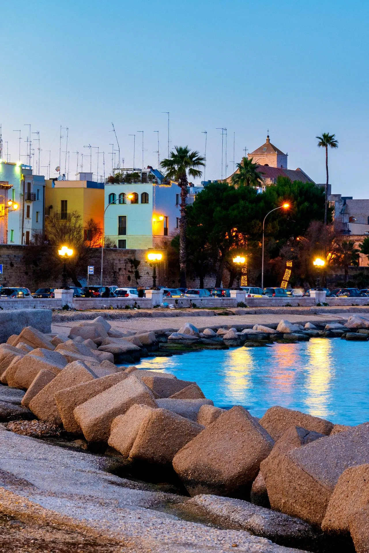 "Schilderachtige kustlijn van Puglia, Italië, met kleurrijke huizen, palmbomen en verlichte straatlantaarns langs de boulevard bij zonsondergang. De Adriatische Zee weerspiegelt het warme licht, terwijl robuuste stenen blokken de kust beschermen. Ideaal voor een zomervakantie in Zuid-Italië, geniet van stranden, historische steden, mediterrane cultuur en lokale gastronomie in deze prachtige regio."