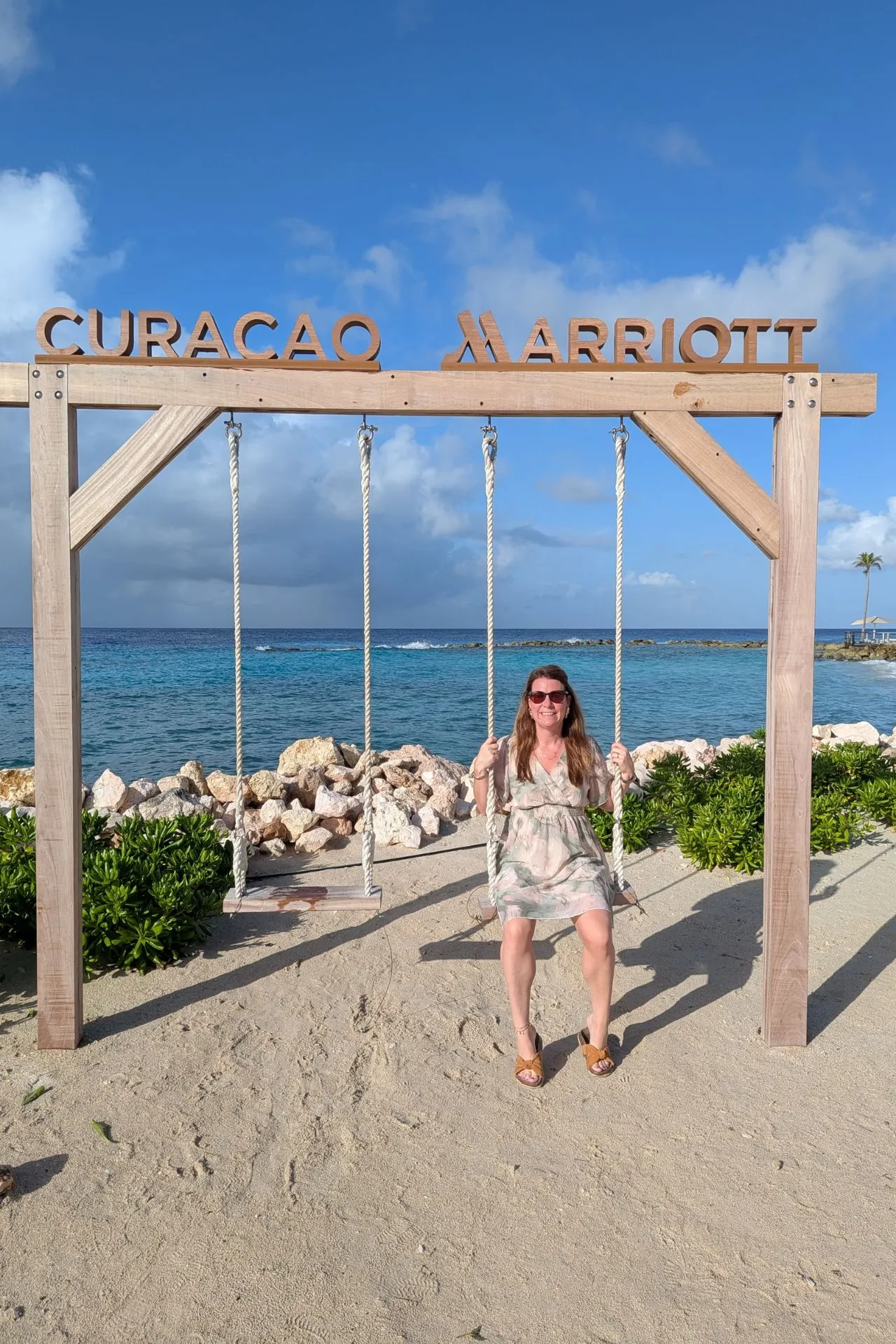 Houten schommel met opschrift Curaçao Marriott op een wit zandstrand met helderblauw water en rotsen op de achtergrond. Tropische sfeer met palmbomen en rustige branding, ideaal voor ontspanning en foto’s tijdens een luxe vakantie op Curaçao. Perfect voor romantische momenten en social media content.