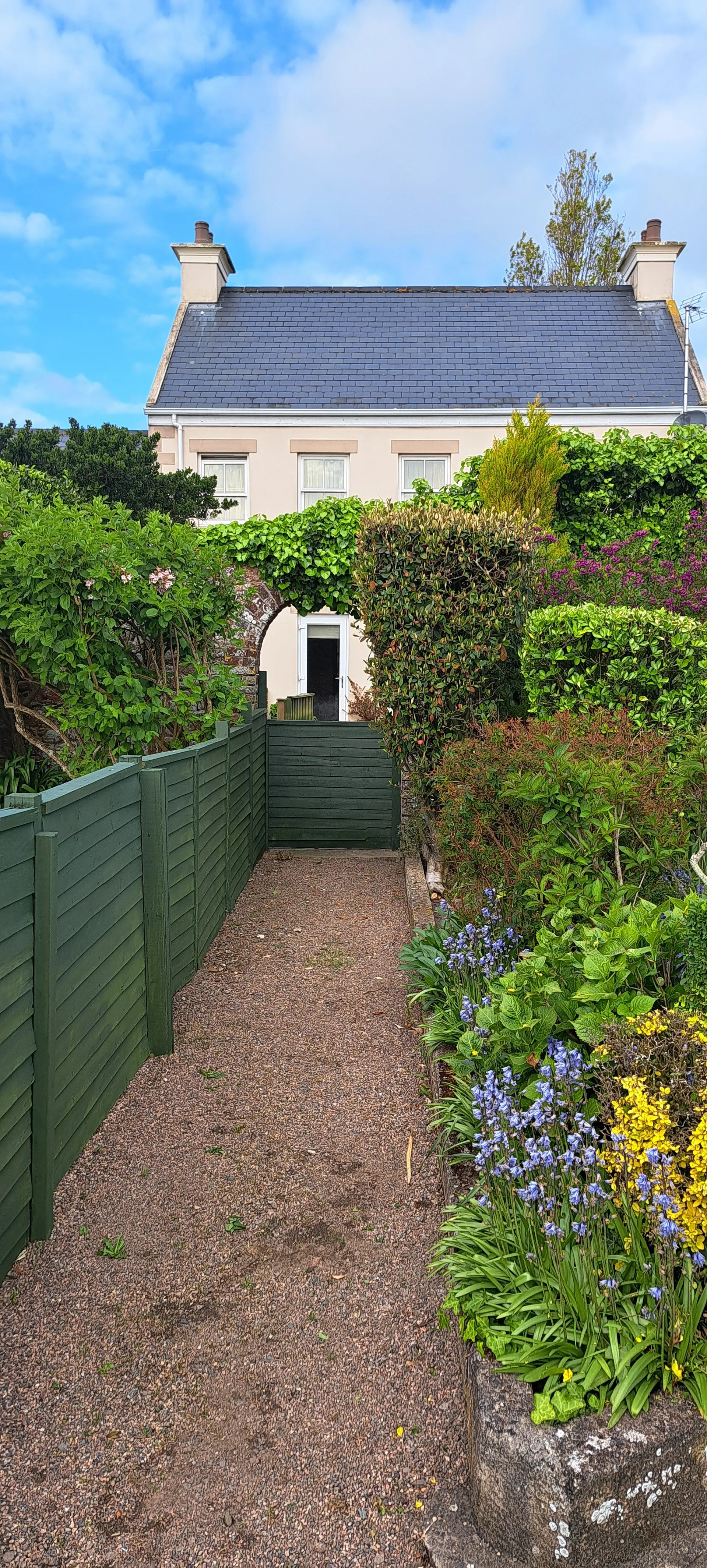Charmante tuinpad met groene houten schutting en kleurrijke bloemen zoals paarse, gele en roze bloesems, leidend naar een traditioneel huis met lichtgekleurde gevel en donkerblauw dak. De tuin is weelderig en goed onderhouden, omringd door struiken en bomen onder een heldere blauwe lucht met lichte wolken. Perfect voor landelijke vakanties, pittoreske accommodaties en natuurbeleving in een rustige omgeving.