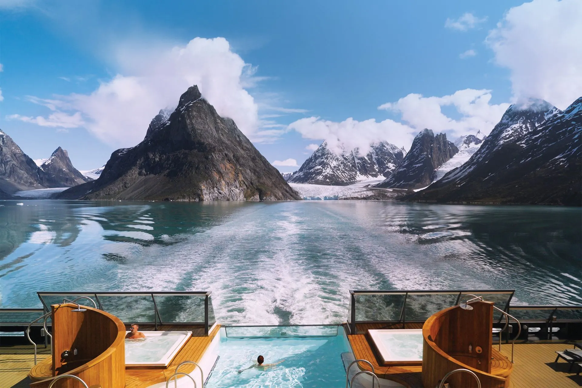 Uitzicht vanaf het achterdek van een Holland America Line cruiseschip met zwembaden en whirlpools, varend door een Noors fjordlandschap met besneeuwde bergtoppen.

