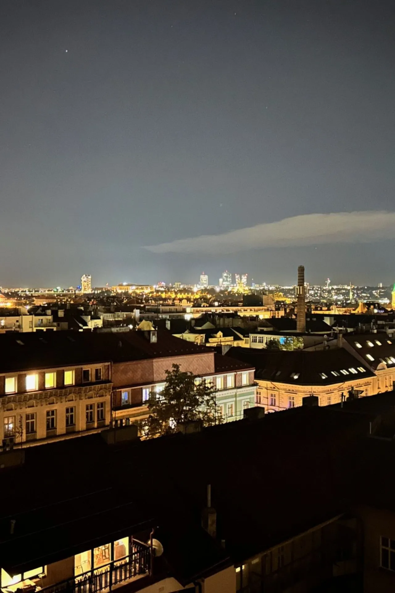 Spectaculair uitzicht over een Europese stad bij nacht, met verlichte gebouwen en een skyline vol moderne torens in de verte. Op de voorgrond zijn klassieke daken en straten zichtbaar, terwijl de horizon fonkelt met stadslichten. De donkere hemel en subtiele wolken creëren een sfeervol contrast. Ideaal voor stedentrips, nachtelijke fotografie en het ontdekken van Praag bij avond.
