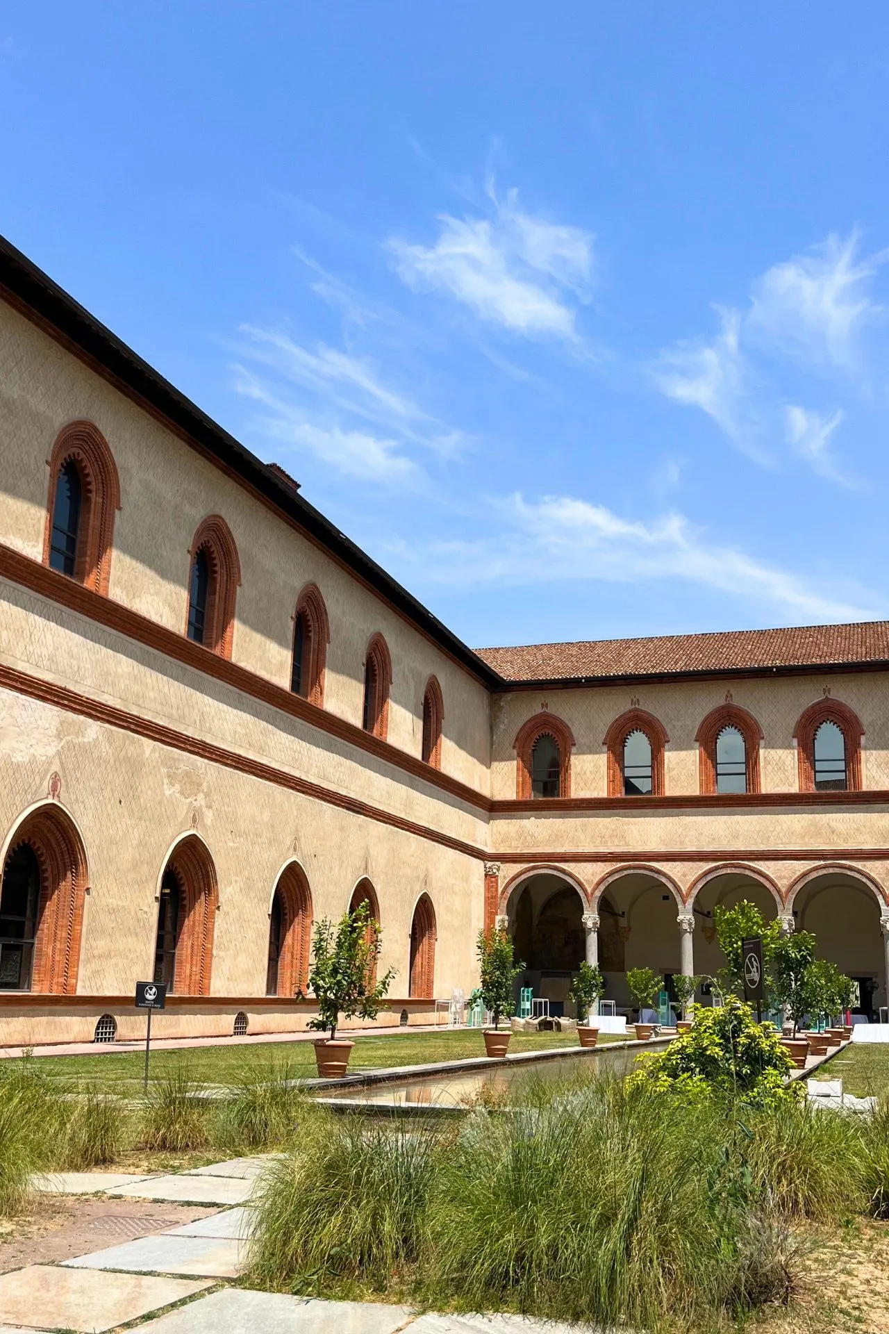 Binnenplaats van het Castello Sforzesco met een verzorgde tuin, zuilengalerij en gotische ramen omlijst met rode baksteen tegen een blauwe lucht.