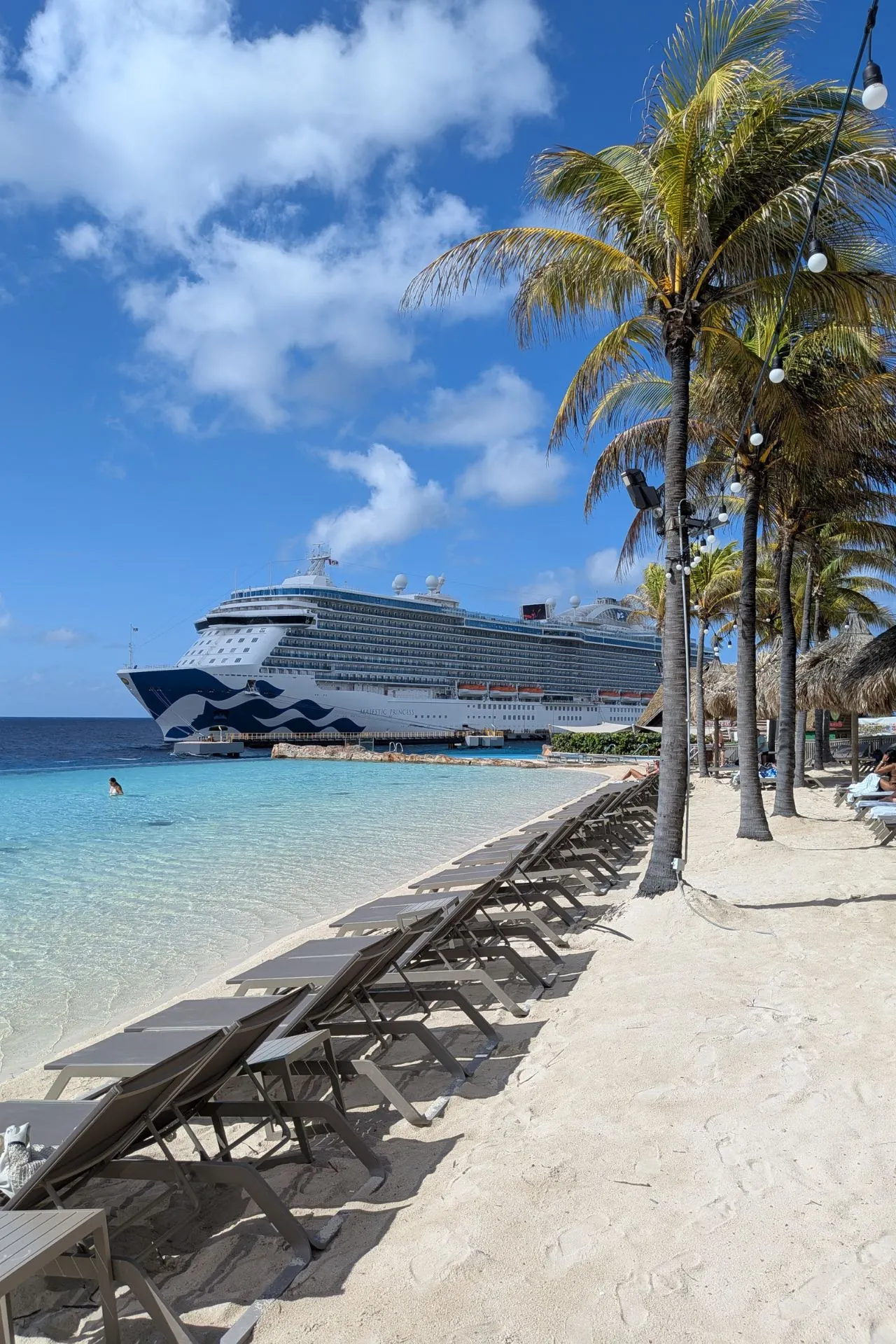 Een idyllisch strand op Curaçao met rijen ligstoelen op het witte zand, palmbomen die zachtjes bewegen in de wind en helder turquoise water dat schittert in de zon. Op de achtergrond ligt een groot cruiseschip aangemeerd, wat een indrukwekkend contrast vormt met de rustige kustlijn. Deze tropische setting straalt ontspanning en luxe uit, perfect voor een zonnige vakantie op het eiland.