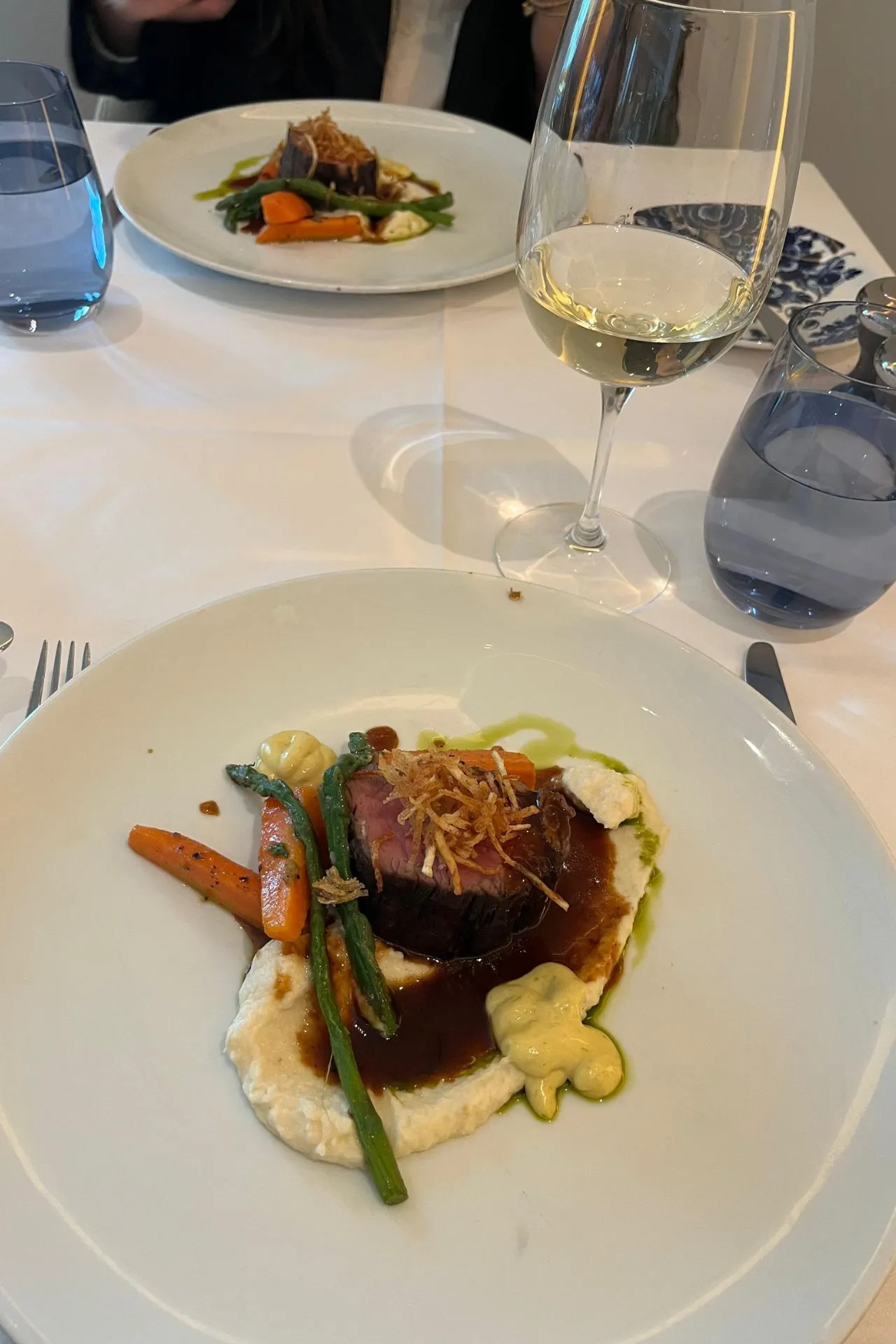 Een elegant gepresenteerd gerecht met groenten en vlees, geserveerd op een witte porseleinen schaal, met een glas witte wijn erbij.