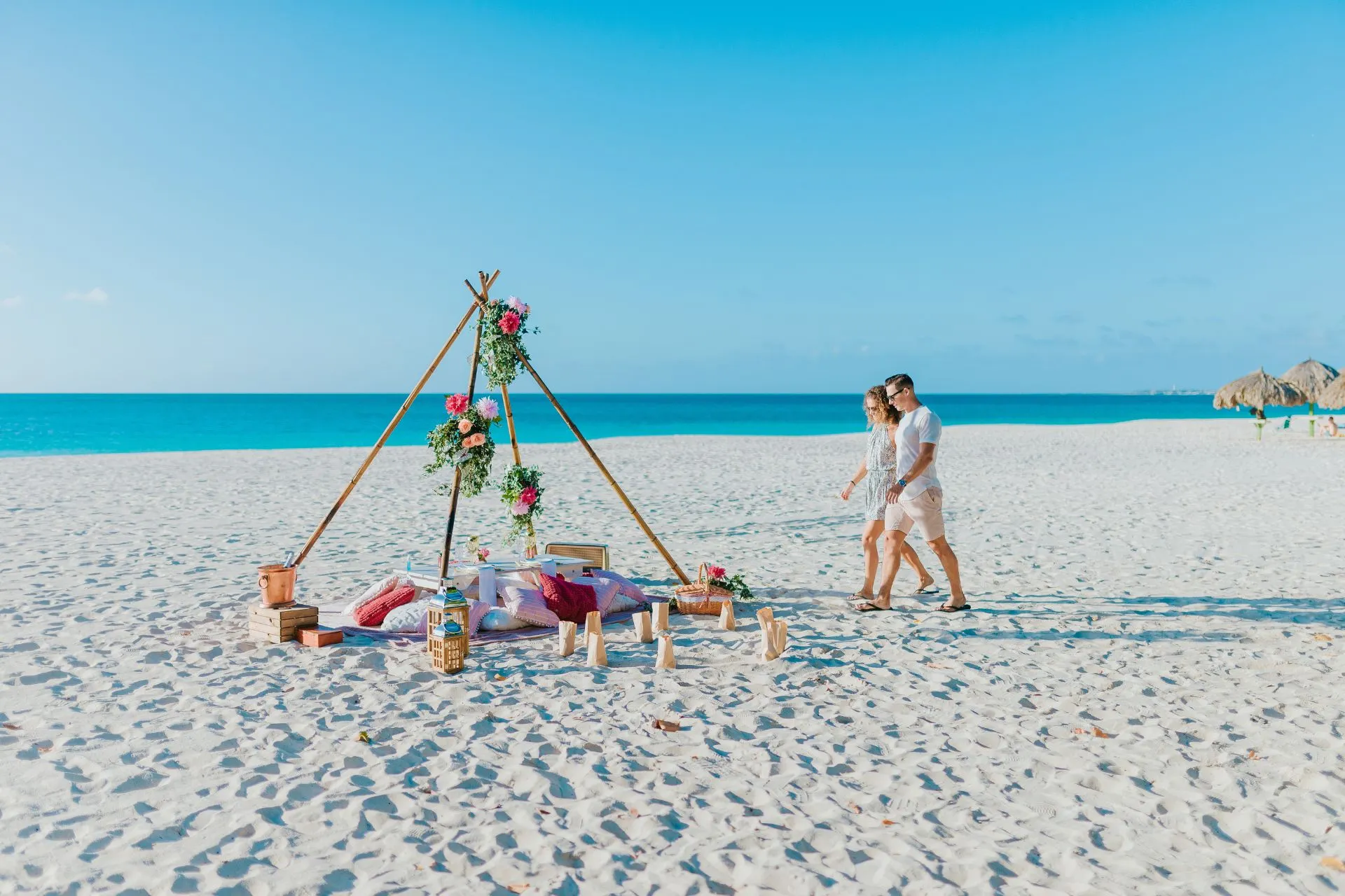 Een idyllisch wit zandstrand met een helderblauwe lucht en een rustige, glinsterende zee op de achtergrond. In het midden staat een romantische picknickopstelling met kaarsen, bloemen en lekkernijen, prachtig uitgestald onder de stralende zon. Een stel loopt hand in hand naar deze uitnodigende setting toe, klaar voor een bijzonder moment samen.
