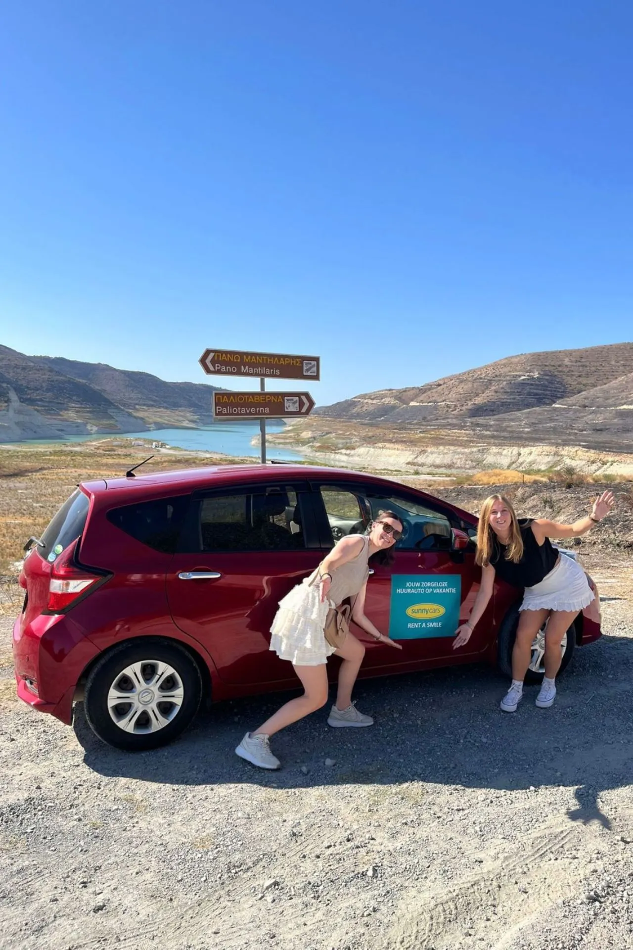 Twee personen poseren naast een rode huurauto van Sunny Cars op een stoffige weg in Cyprus, met wegwijzers en een meer op de achtergrond.