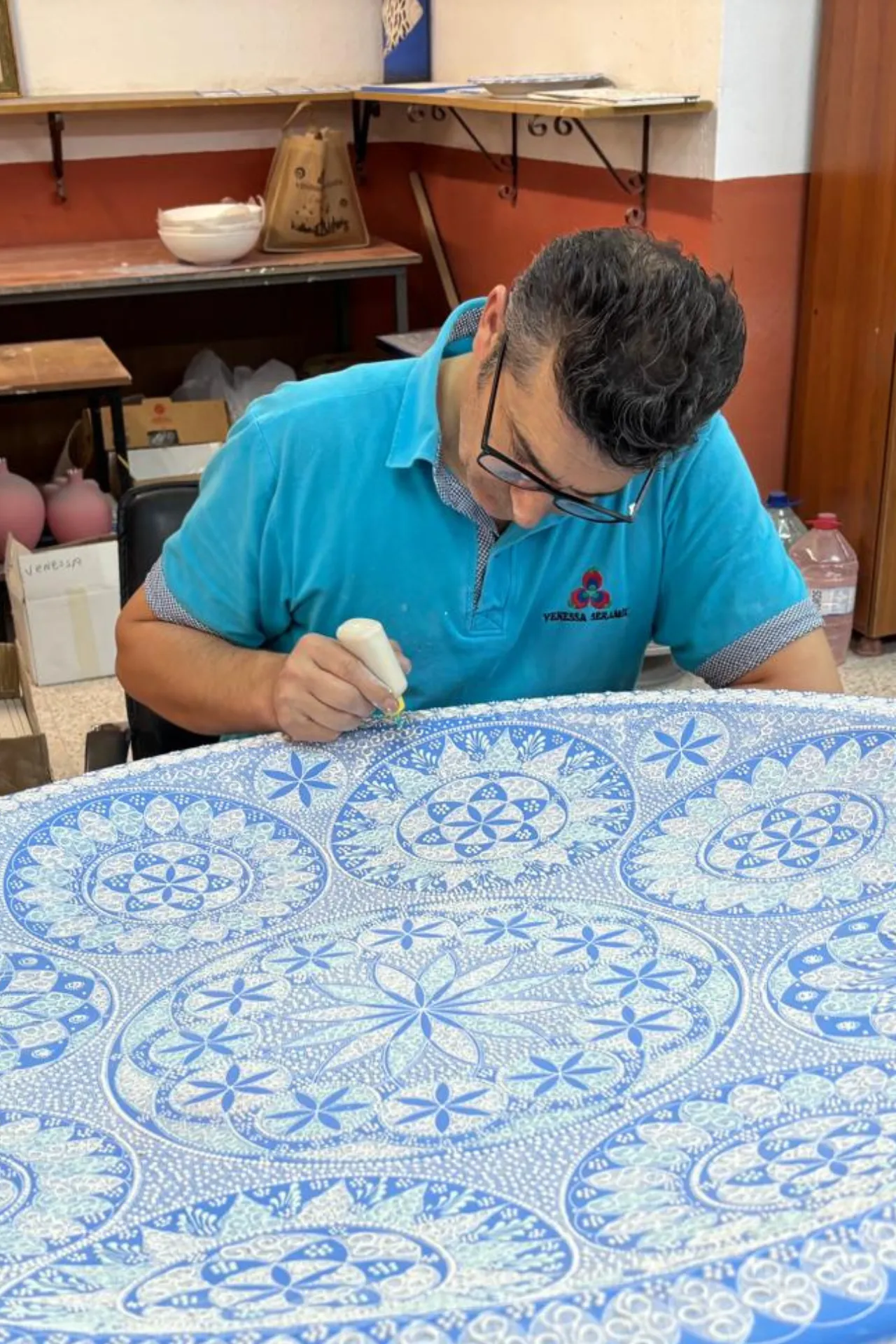 Ambachtsman werkt aan een groot keramisch kunstwerk in Avanos, Cappadocië, Turkije. Het ronde oppervlak is gedecoreerd met ingewikkelde blauwe patronen en traditionele motieven, zorgvuldig aangebracht met een fijne penseeltechniek. De werkruimte op de achtergrond toont potten, gereedschap en aardetinten die de authentieke sfeer van een pottenbakkerij benadrukken. Deze ambachtelijke kunstvorm is een belangrijk onderdeel van de Turkse cultuur en trekt toeristen en liefhebbers van handgemaakte keramiek en design.