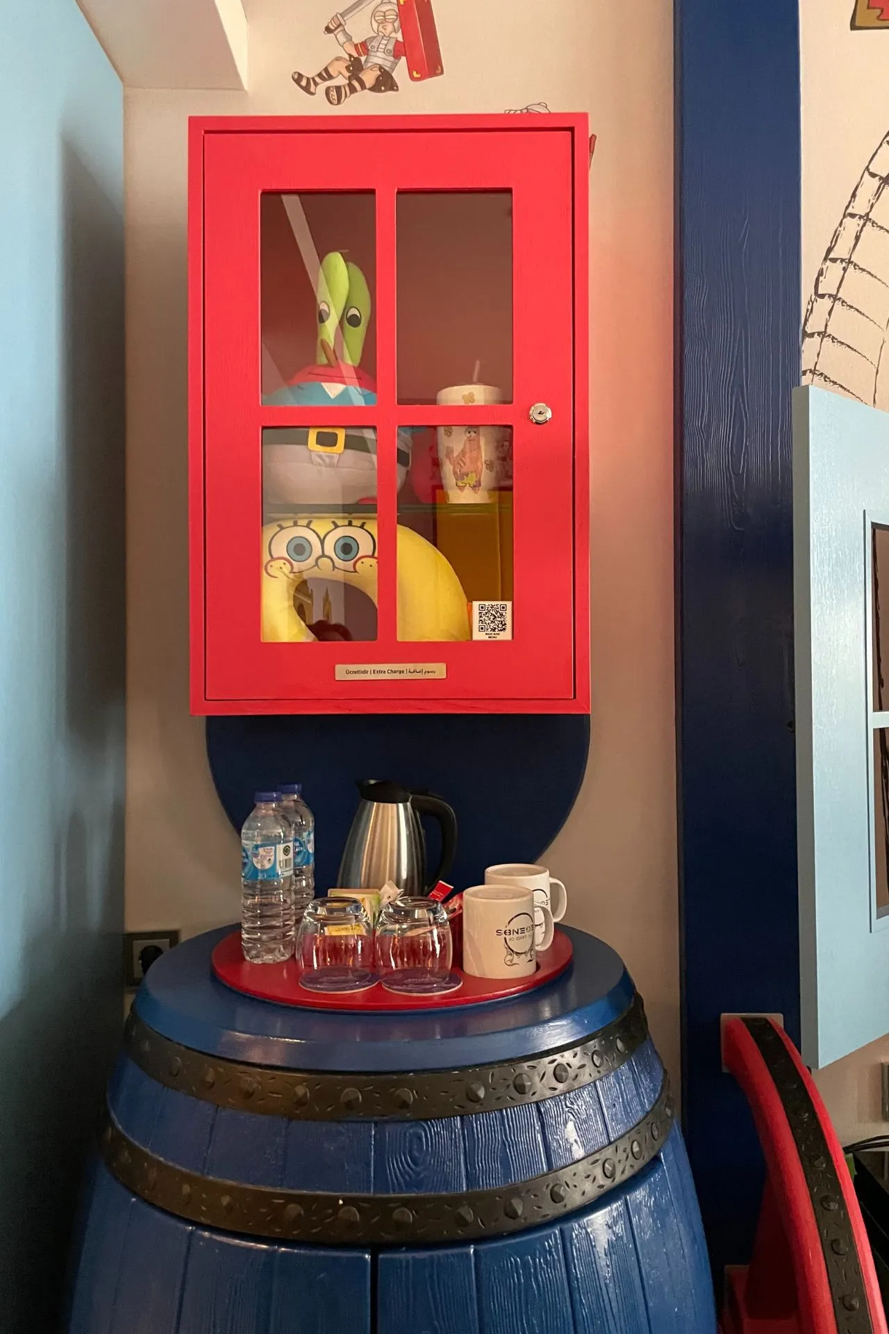 Minibarhoek met een rode kast waarin een SpongeBob-figuur zichtbaar is, naast een blauwe ton met glazen en een waterfles.