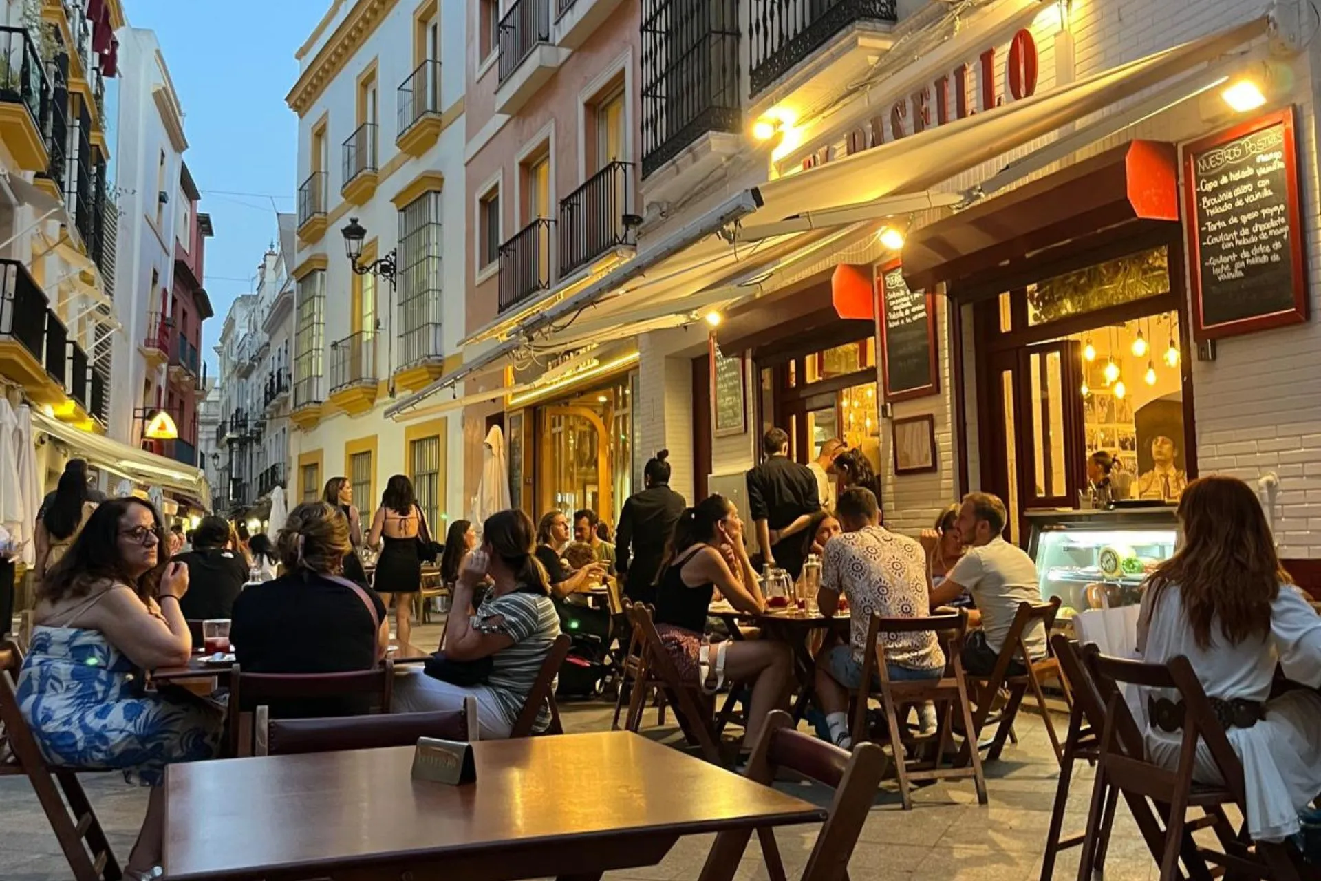 Gezellige drukte op het terras van El Paseillo in Sevilla tijdens de avondschemering. Mensen genieten van eten en drankjes aan houten tafeltjes in een smalle straat met kleurrijke gevels en sfeervolle verlichting.


