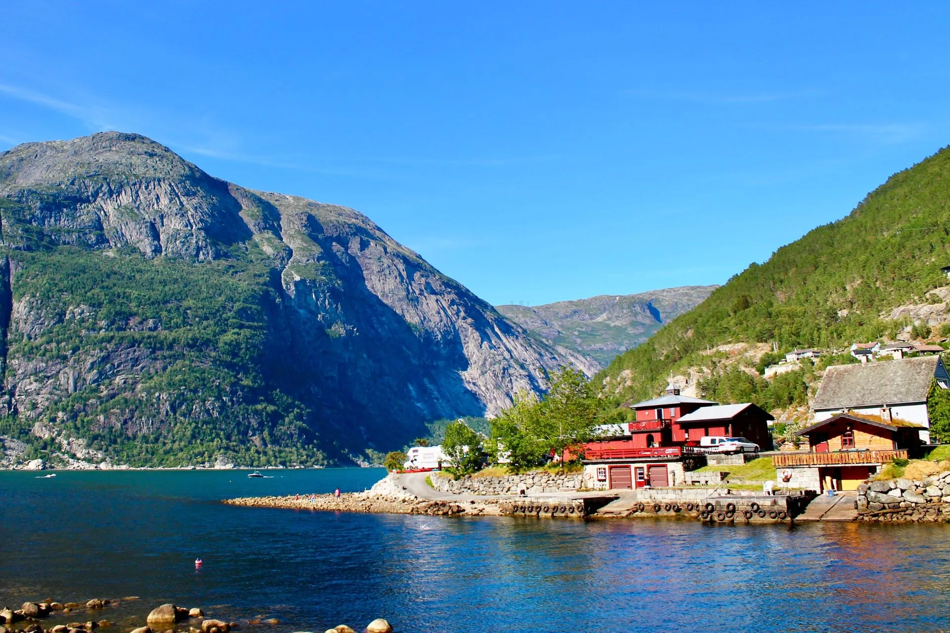 Uitzicht op een fjord in Noorwegen met rode huisjes aan het water, omringd door hoge bergwanden en groene bossen onder een stralend blauwe lucht.
