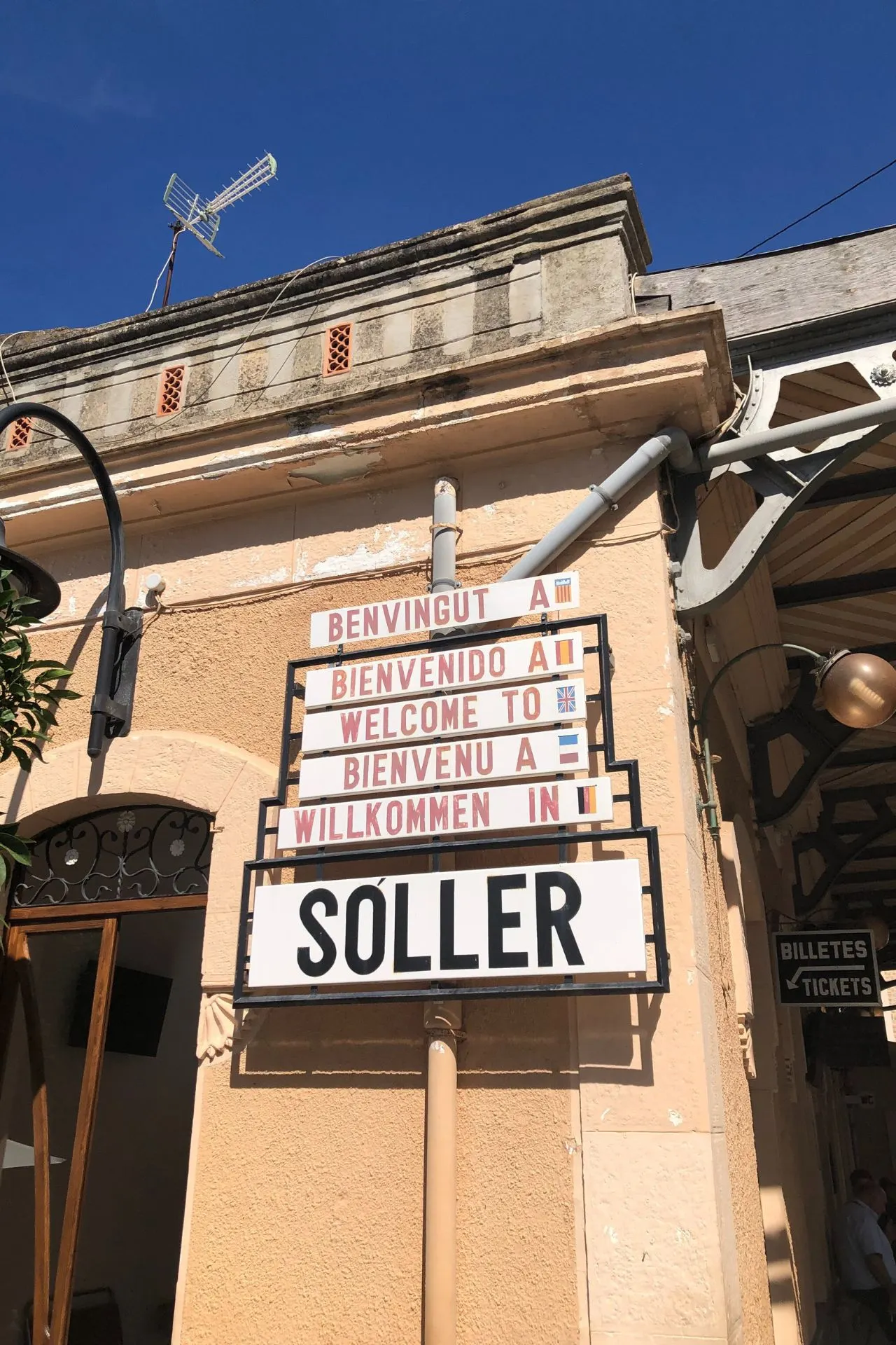 Welkomstbord bij het treinstation van Sóller op Mallorca met tekst in meerdere talen, waaronder Spaans, Engels en Duits, onder een blauwe lucht. Sóller is beroemd om zijn nostalgische houten trein die door de bergen naar Palma de Mallorca rijdt. Ideaal voor toeristen die een authentieke reiservaring en prachtige uitzichten willen beleven tijdens hun vakantie op Mallorca.
