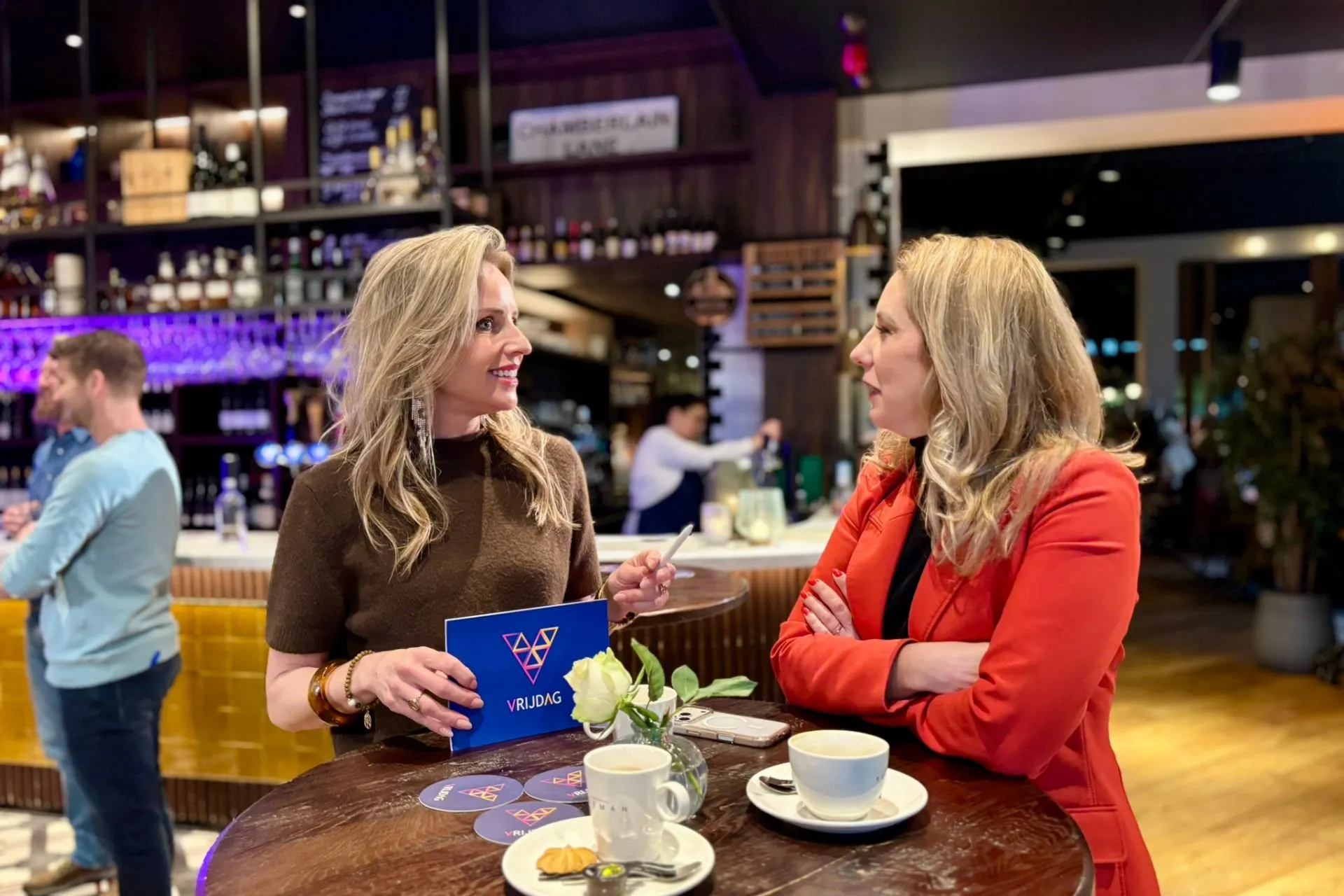 Twee personen in een levendige baromgeving voeren een gesprek aan een ronde houten tafel met koffie, een witte roos en een opvallend blauw kaartje met het woord “Vrijdag”. Op de achtergrond een goed gevulde bar met flessen en glazen, wat zorgt voor een stijlvolle en uitnodigende sfeer. Deze setting benadrukt het belang van persoonlijke interactie tijdens zakelijke borrels, netwerkbijeenkomsten en informele ontmoetingen in een moderne horecalocatie met warme verlichting.
