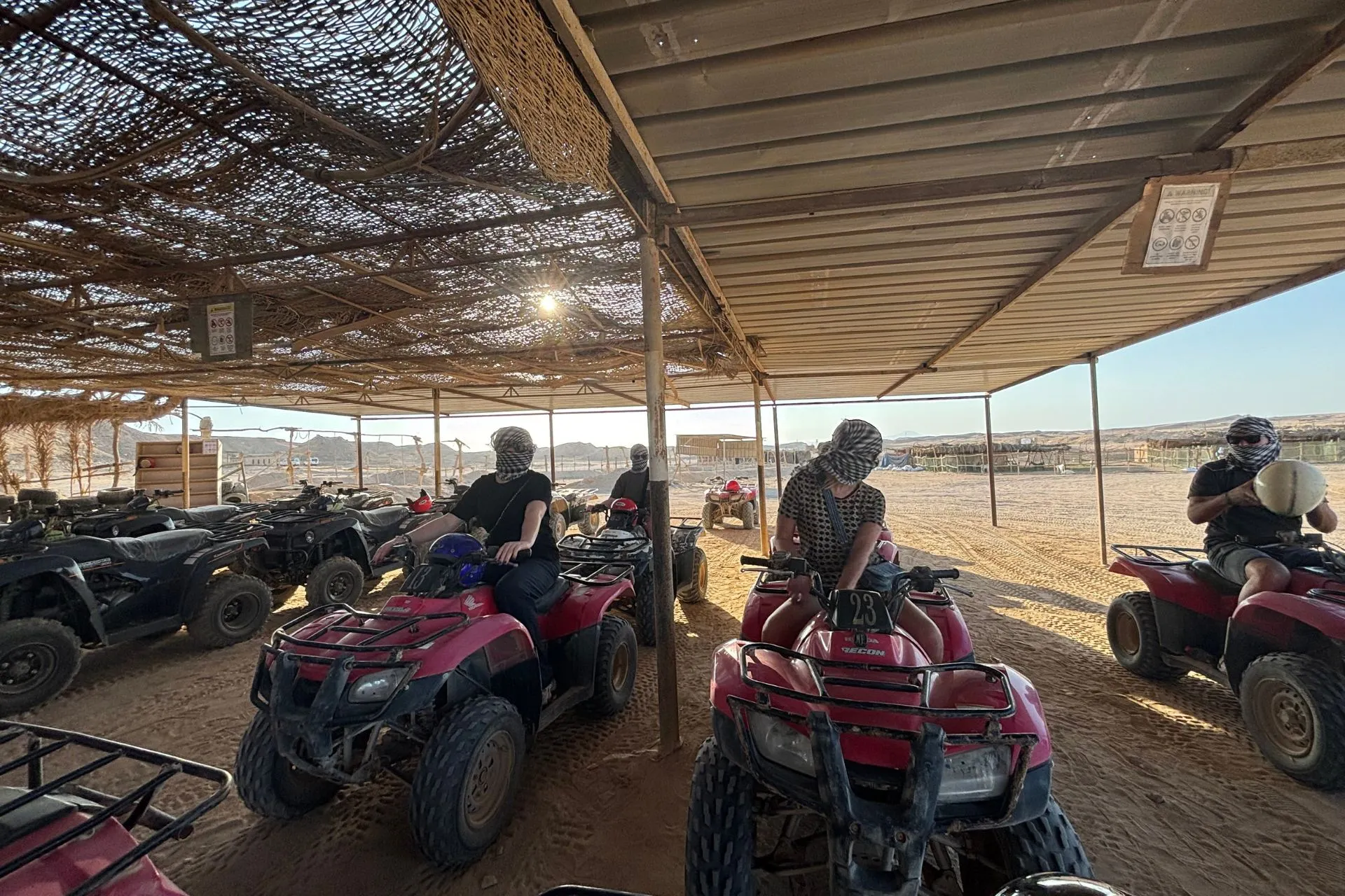 Meerdere rode quads staan opgesteld onder een schaduwrijke overkapping van gevlochten materiaal in een woestijnkamp. Mensen zitten op de quads met helmen en sjaals om hun gezicht te beschermen tegen stof, klaar voor vertrek. Op de achtergrond is een open woestijnlandschap zichtbaar met zand, lage heuvels en een heldere lucht. Dit beeld benadrukt avontuur en voorbereiding voor een quadtocht, ideaal voor promotie van woestijnexcursies, actieve vakanties en outdoor activiteiten in Egypte.