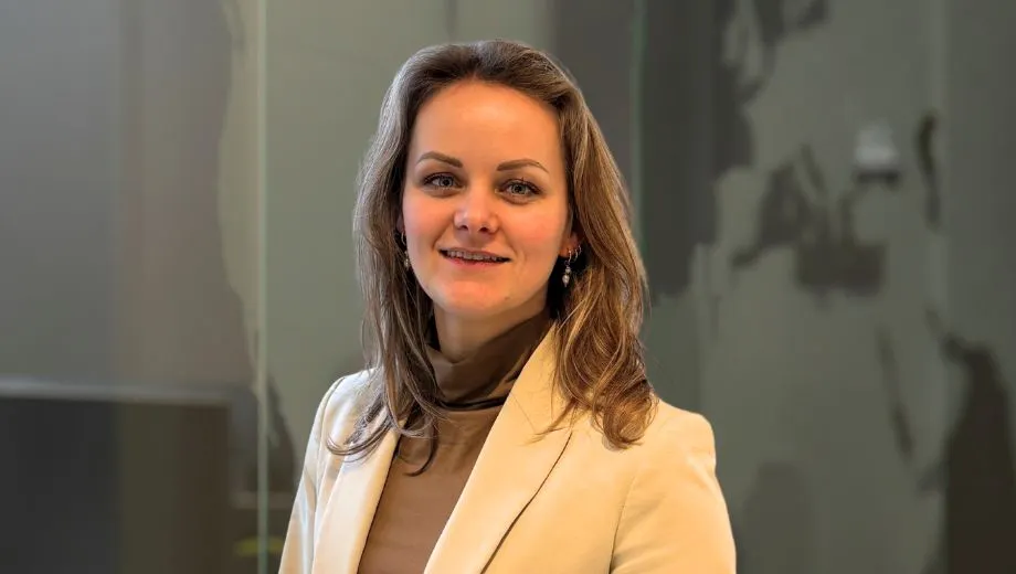De foto toont Manon onze creatieve marketeer in een professionele setting, gekleed in een zwarte blazer.Het haar is blond van kleur en valt in losse krullen over de schouders. De achtergrond bestaat uit een moderne kantooromgeving met glazen wanden en een subtiele reflectie, wat een zakelijke en formele sfeer creëert. De verlichting is helder en neutraal, waardoor de details van de kleding en omgeving goed zichtbaar zijn.