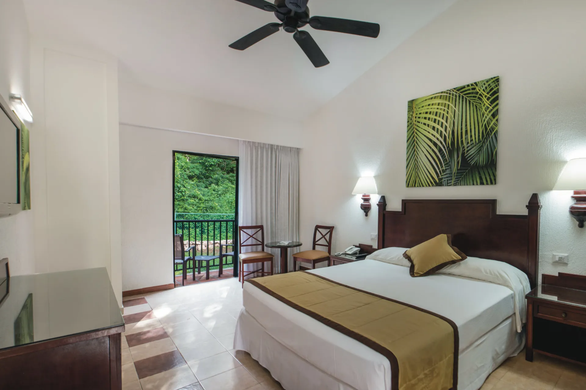 Een lichte en ruime hotelkamer met een groot tweepersoonsbed, voorzien van witte lakens en een beige bedloper. Aan weerszijden van het bed staan houten nachtkastjes met lampen, en boven het bed hangt een kunstwerk met groene palmbladeren. De kamer heeft een plafondventilator en een tegelvloer in neutrale tinten. Aan de achterzijde bevindt zich een glazen schuifdeur die leidt naar een balkon met uitzicht op weelderige groene natuur, wat een rustige en tropische sfeer creëert.