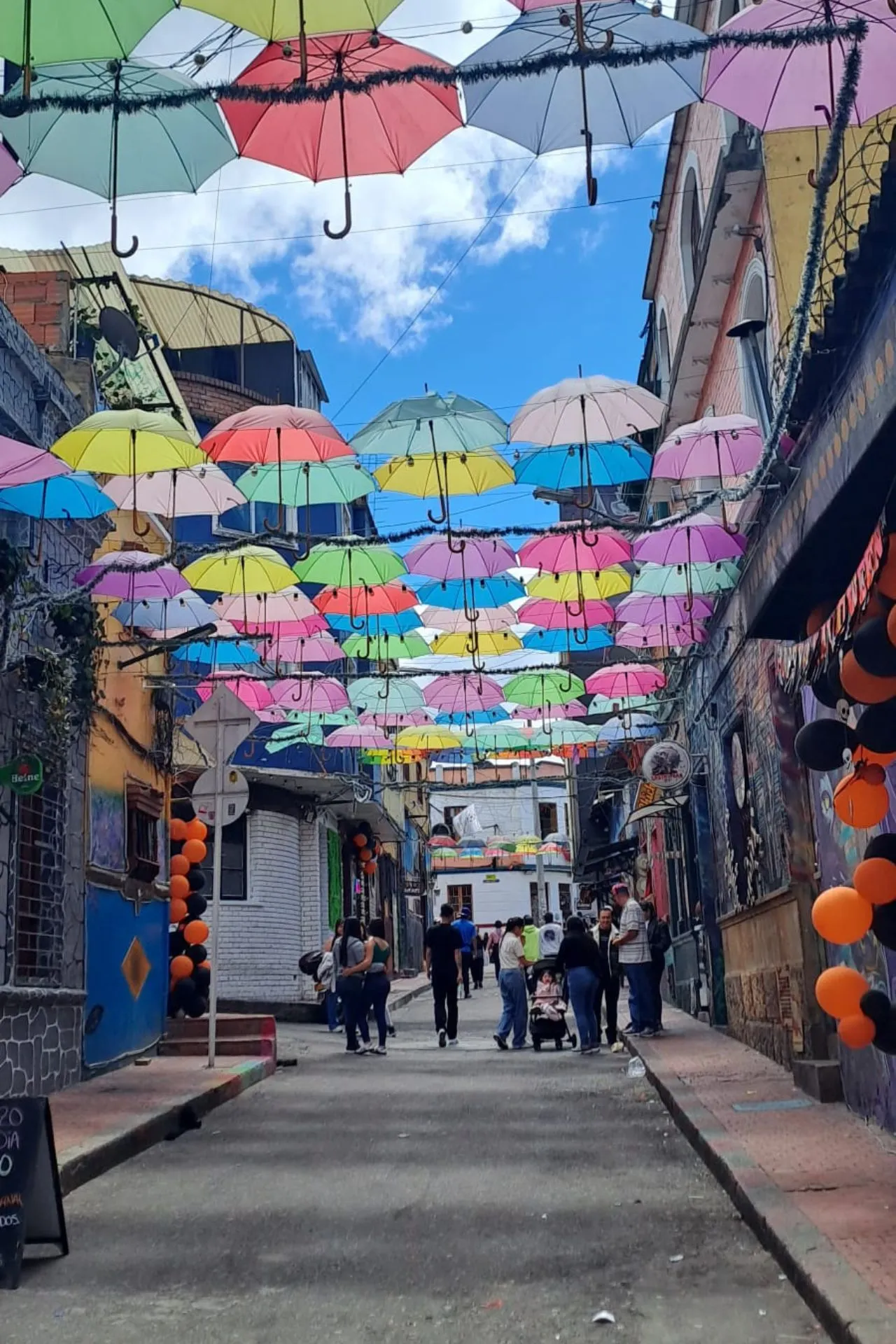Straat met een plafond van kleurrijke paraplu’s in roze, blauw, groen en geel, met mensen die door de steeg lopen.