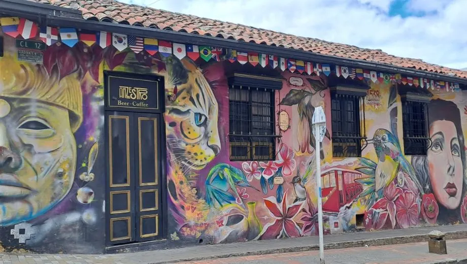 Artistieke muurschildering op een gebouw met afbeeldingen van een jaguar, vogels, bloemen en menselijke gezichten, onder een rij internationale vlaggen.