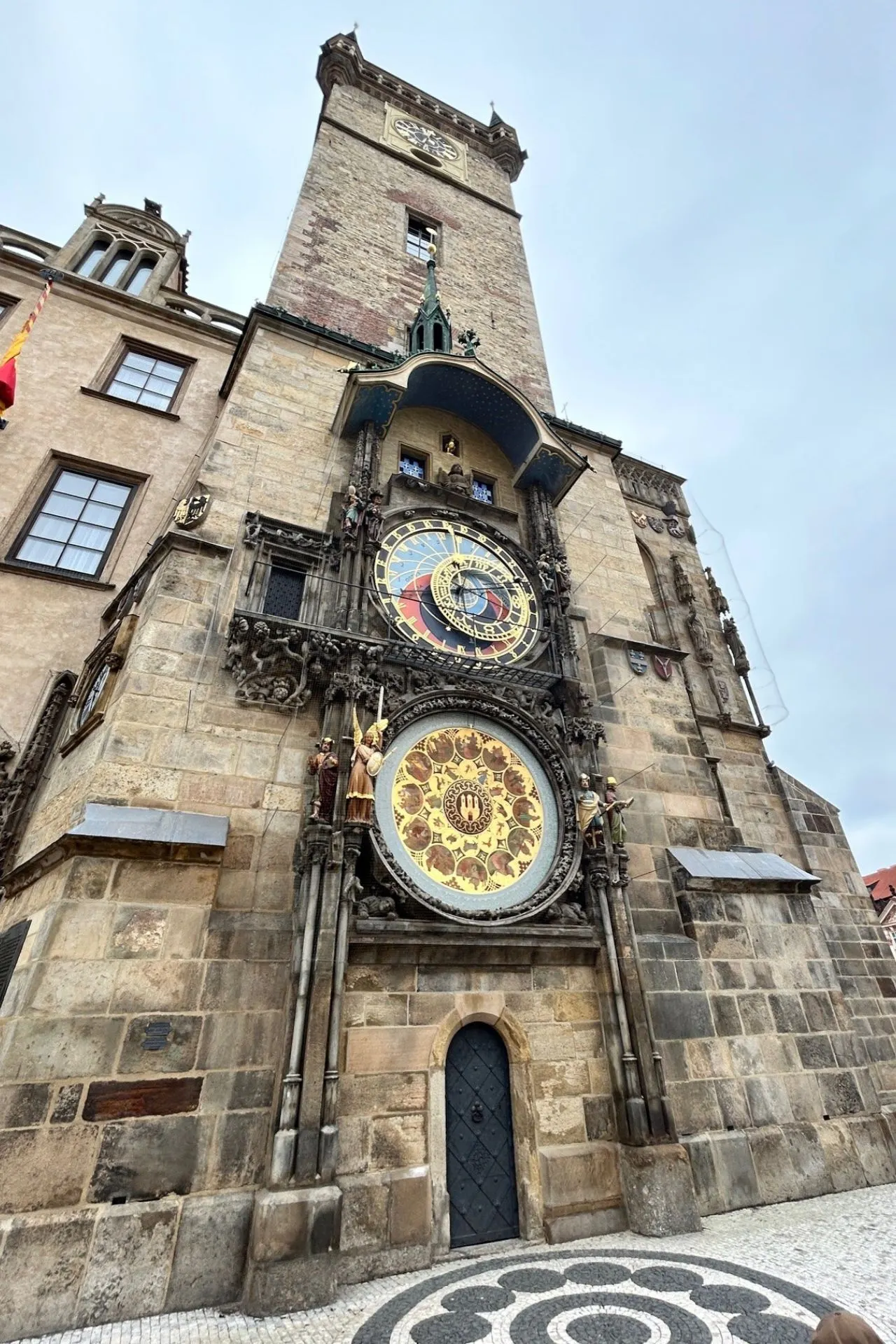 Close-up van de beroemde astronomische klok op een historische toren, met gedetailleerde gouden en kleurrijke wijzerplaten en decoratieve beelden. De toren is gebouwd van natuursteen en heeft gotische elementen en een imposante hoogte. De geplaveide straat en het patroon op de grond versterken de authentieke sfeer. Iconische bezienswaardigheid voor toeristen en liefhebbers van geschiedenis en architectuur.

