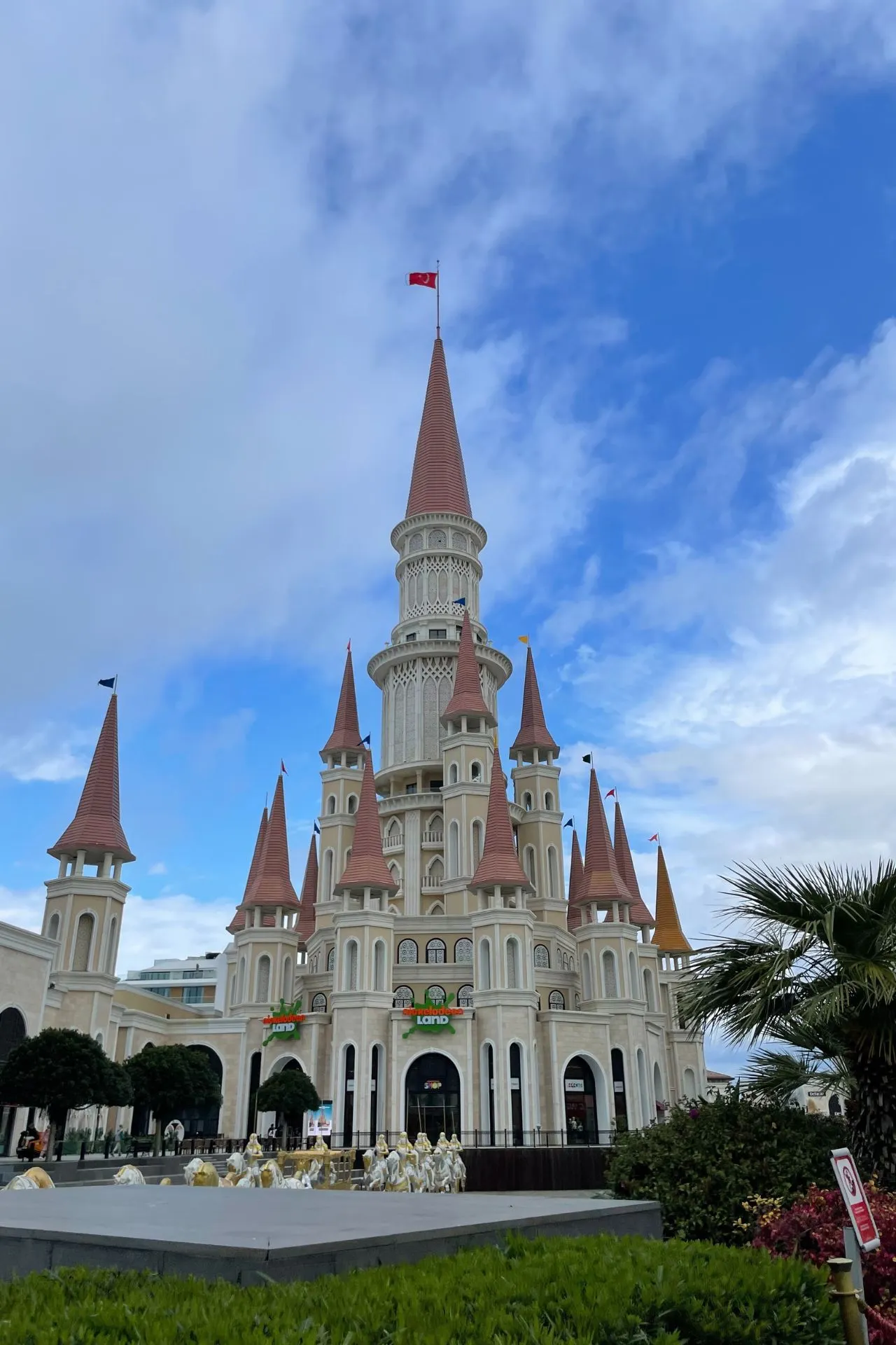 Groot sprookjesachtig kasteel met meerdere torens en rode puntdaken, bekroond met een rode vlag bovenop de hoogste toren. Het gebouw heeft witte gevels met sierlijke details en staat tegen een blauwe lucht met enkele wolken. Op de voorgrond zijn groene struiken en palmbomen zichtbaar.