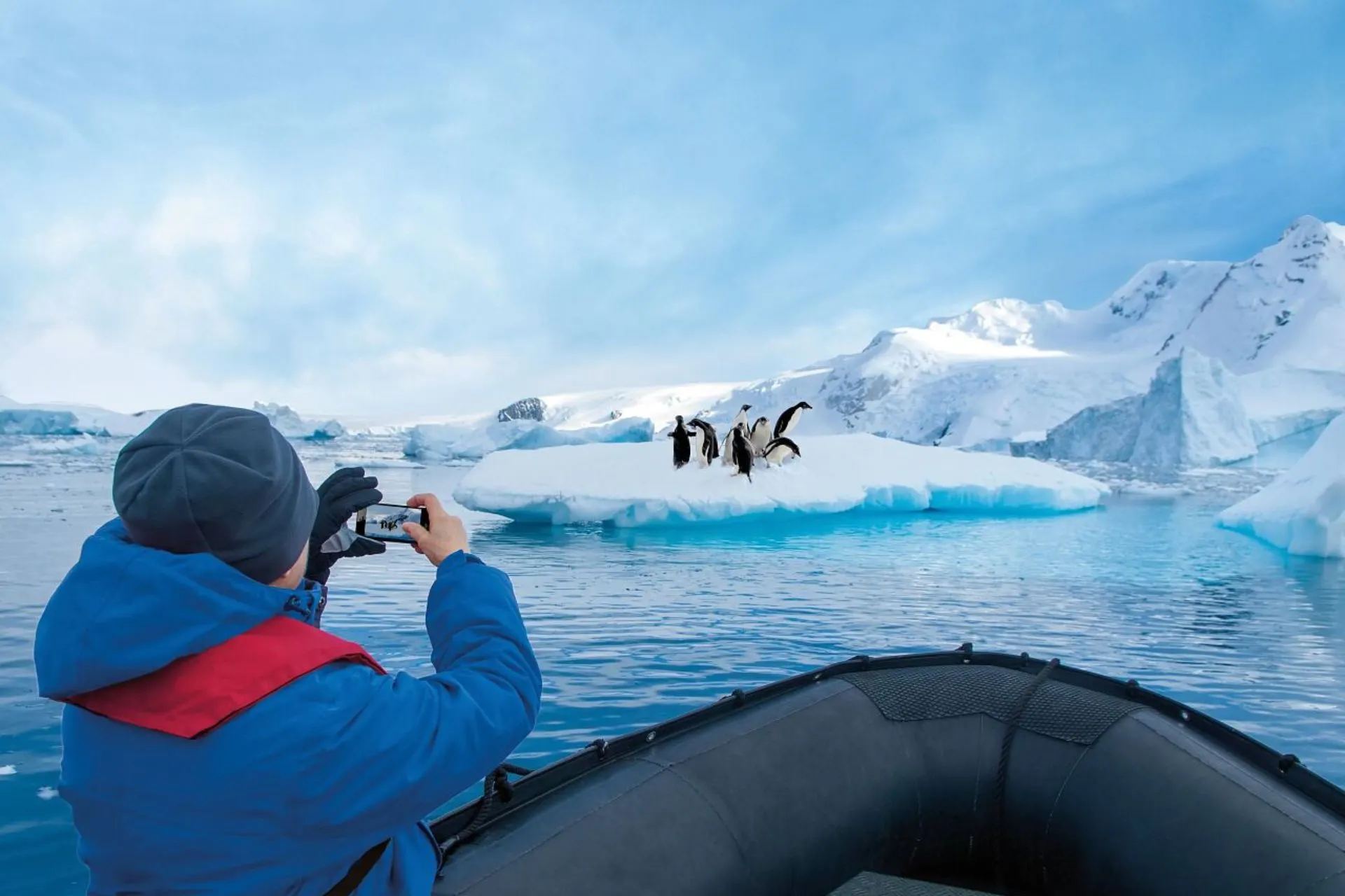Passagier van een Hapag-Lloyd expeditiecruise maakt een foto van pinguïns op een ijsschots tijdens een zodiacexcursie in het indrukwekkende landschap van Antarctica, omringd door ijsbergen en besneeuwde bergen.