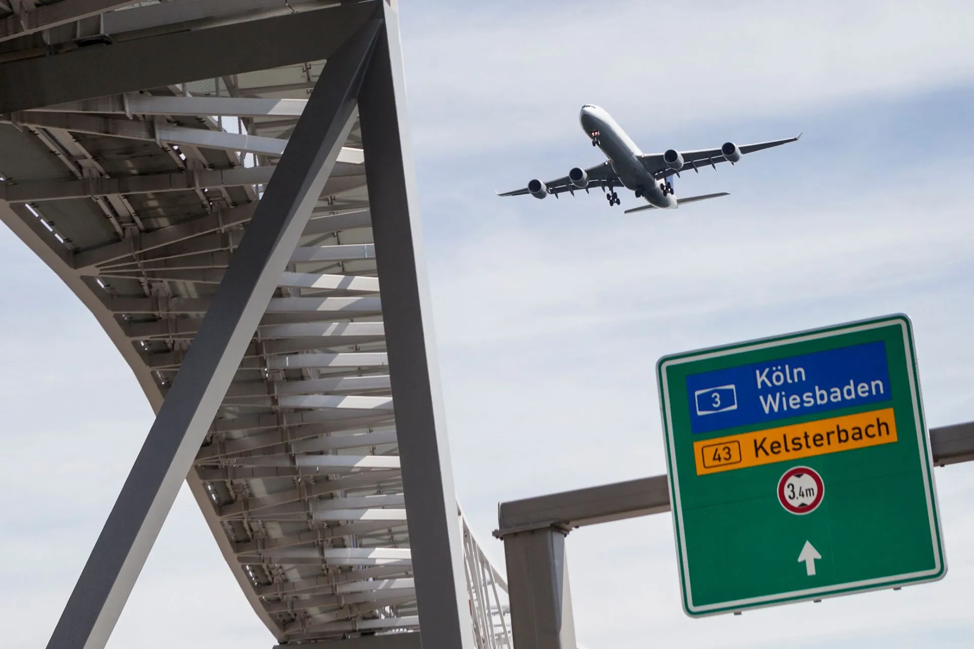 Vliegtuig dat over een snelweg vliegt bij luchthaven Keulen Bonn, met verkeersborden naar Keulen en Wiesbaden. Perfect voor vertrek vanaf luchthaven Keulen.
