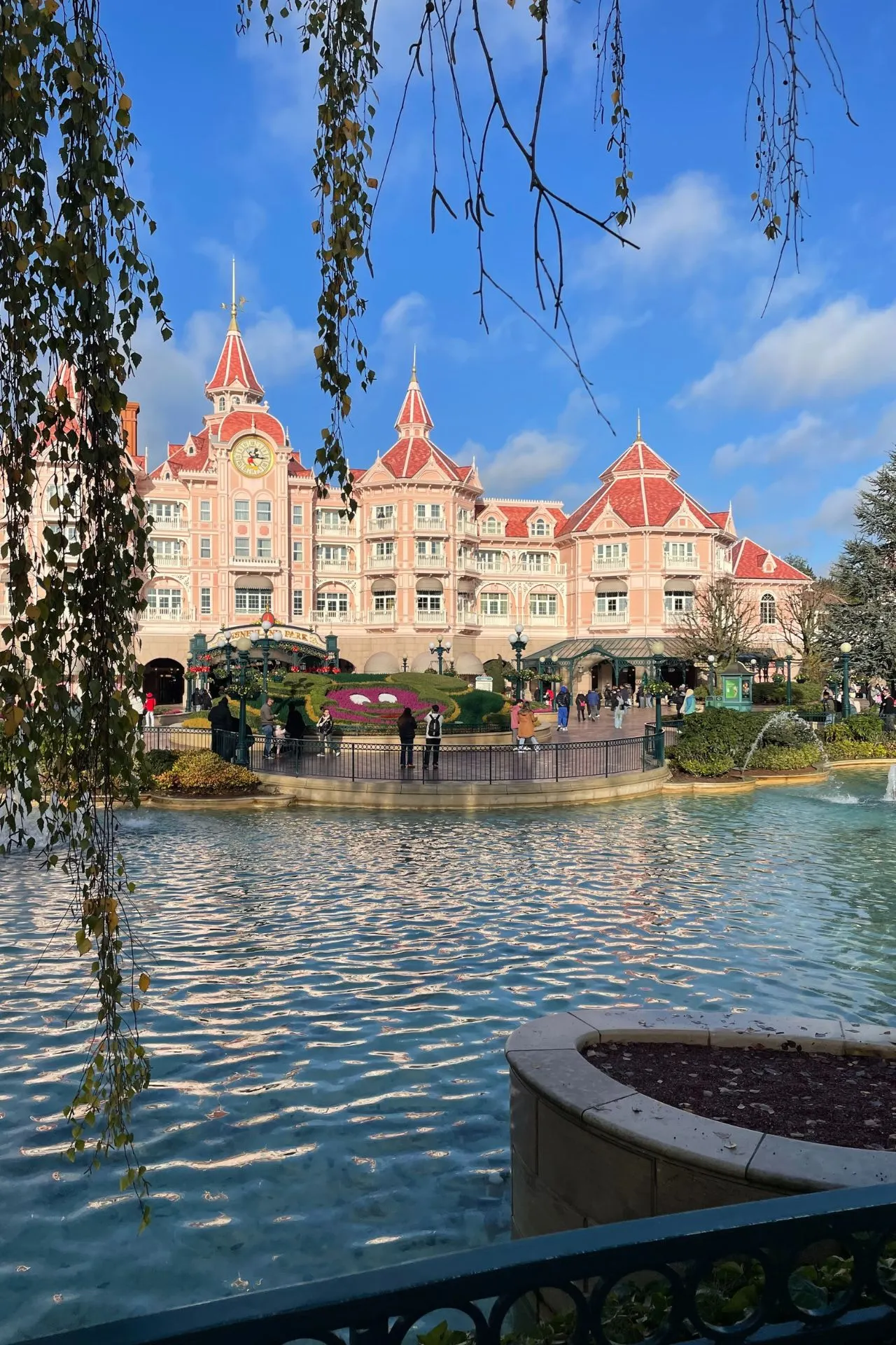 Het iconische roze Disneyland Hotel in Disneyland Parijs weerspiegelt prachtig in het water van de vijver bij de ingang van Disneyland Park. Onder een blauwe lucht wandelen bezoekers richting de hoofdingang van het populaire pretpark. De sprookjesachtige architectuur, romantische torentjes en serene omgeving benadrukken de luxe en magie die veel reizigers zoeken tijdens hun Disneyland Parijs vakantie of Disney trip met overnachting in het Disneyland Hotel.