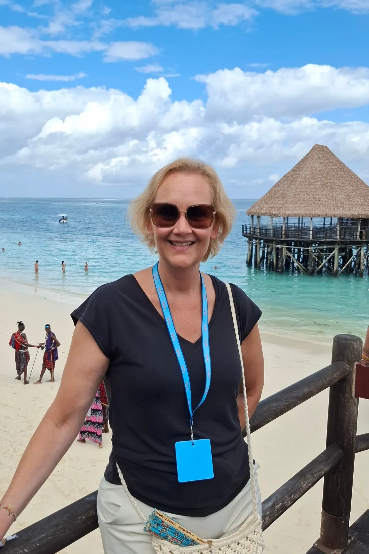 Astrid tijdens een studiereis op Zanzibar bij een wit zandstrand en helderblauwe zee. Op de achtergrond staat een karakteristieke houten pier met rieten dak boven het water. De foto laat de tropische sfeer van Zanzibar zien, met palmbomen, strandactiviteiten en het turquoise water van de Indische Oceaan.