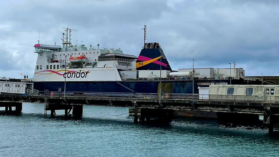 Grote veerboot van Condor Ferries aangemeerd aan een pier, met duidelijk zichtbaar logo en kleurrijke accenten op de schoorsteen. Het schip ligt in helderblauw water onder een bewolkte lucht, wat zorgt voor een contrasterende maritieme sfeer. Op de voorgrond is de aanlegsteiger te zien met voertuigen en infrastructuur voor passagiers en vracht. Deze locatie is ideaal voor reizigers die op zoek zijn naar ferryverbindingen, kusttransport en een authentieke havenervaring.