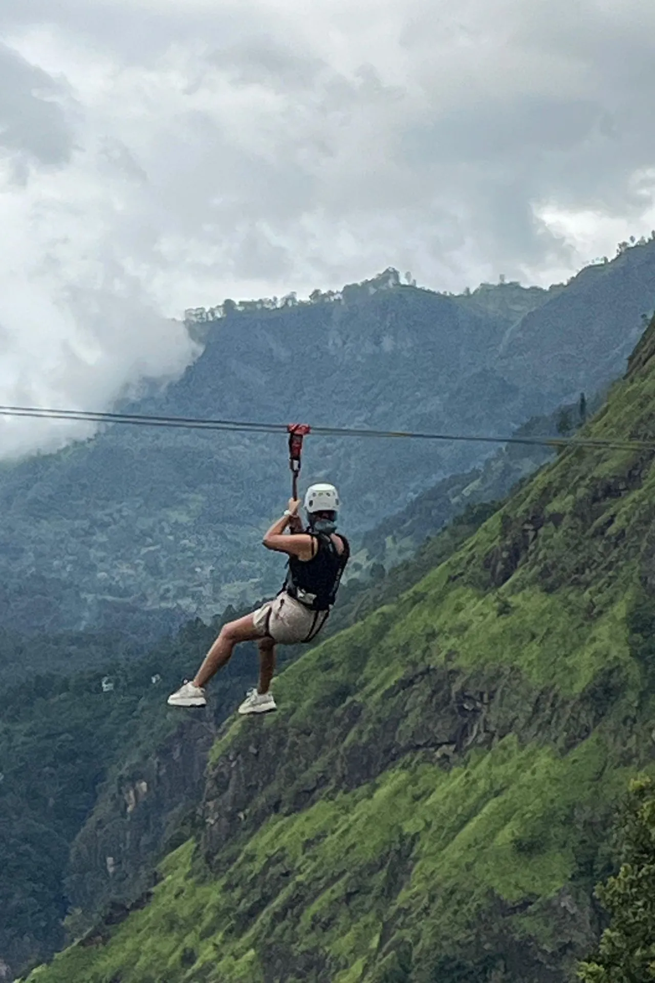 Een avontuurlijke reiziger hangt aan een zipline, hoog boven de groene heuvels van Ella, Sri Lanka. De dramatische berglandschappen en lage wolken op de achtergrond benadrukken de spanning en schoonheid van deze adembenemende rit over de vallei bij Flying Ravana.