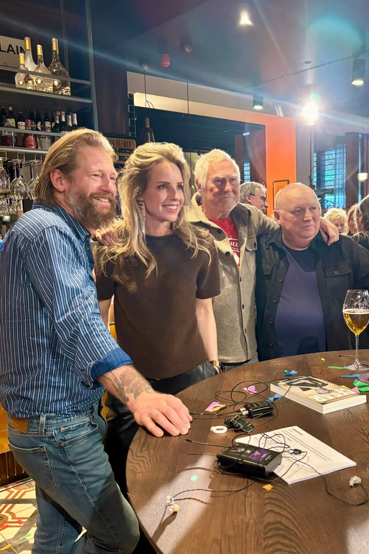 Gezellige bijeenkomst in een moderne bar met een ronde houten tafel vol apparatuur, papieren en kleurrijke confetti. Op de achtergrond een goed gevulde bar met flessen en glazen, terwijl op tafel een glas bier staat. De setting combineert een professionele sfeer met informele gezelligheid, perfect voor bedrijfsborrels, interviews of opnames in een warme en stijlvolle horecalocatie.