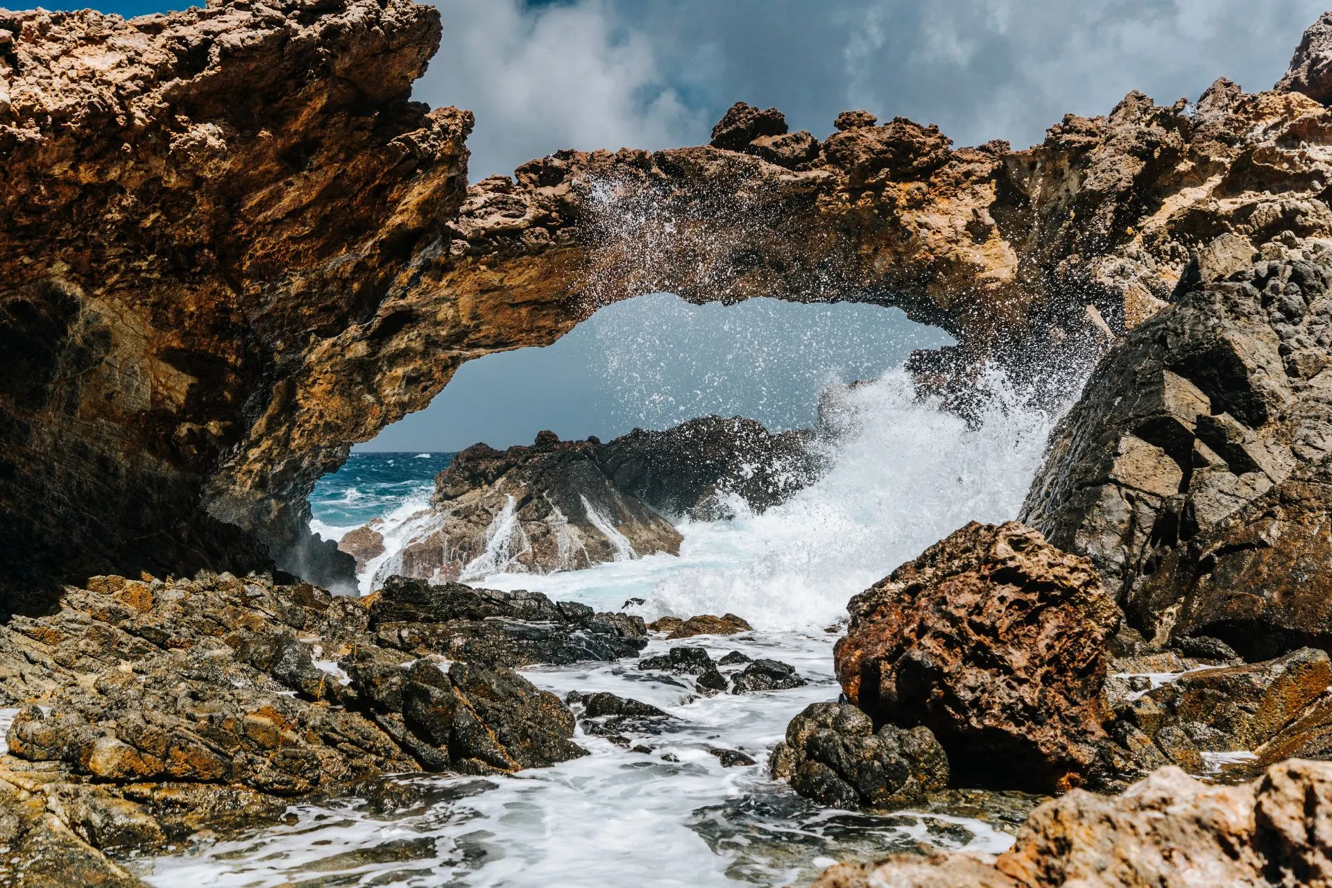 Een indrukwekkend natuurfenomeen op Aruba, waar golven krachtig tegen een grote natuurlijke rotsboog slaan. De rotsen zijn ruw en warmbruin van kleur, terwijl het opspattende water witte schuimkoppen vormt. Op de achtergrond is de diepblauwe zee zichtbaar onder een deels bewolkte lucht.