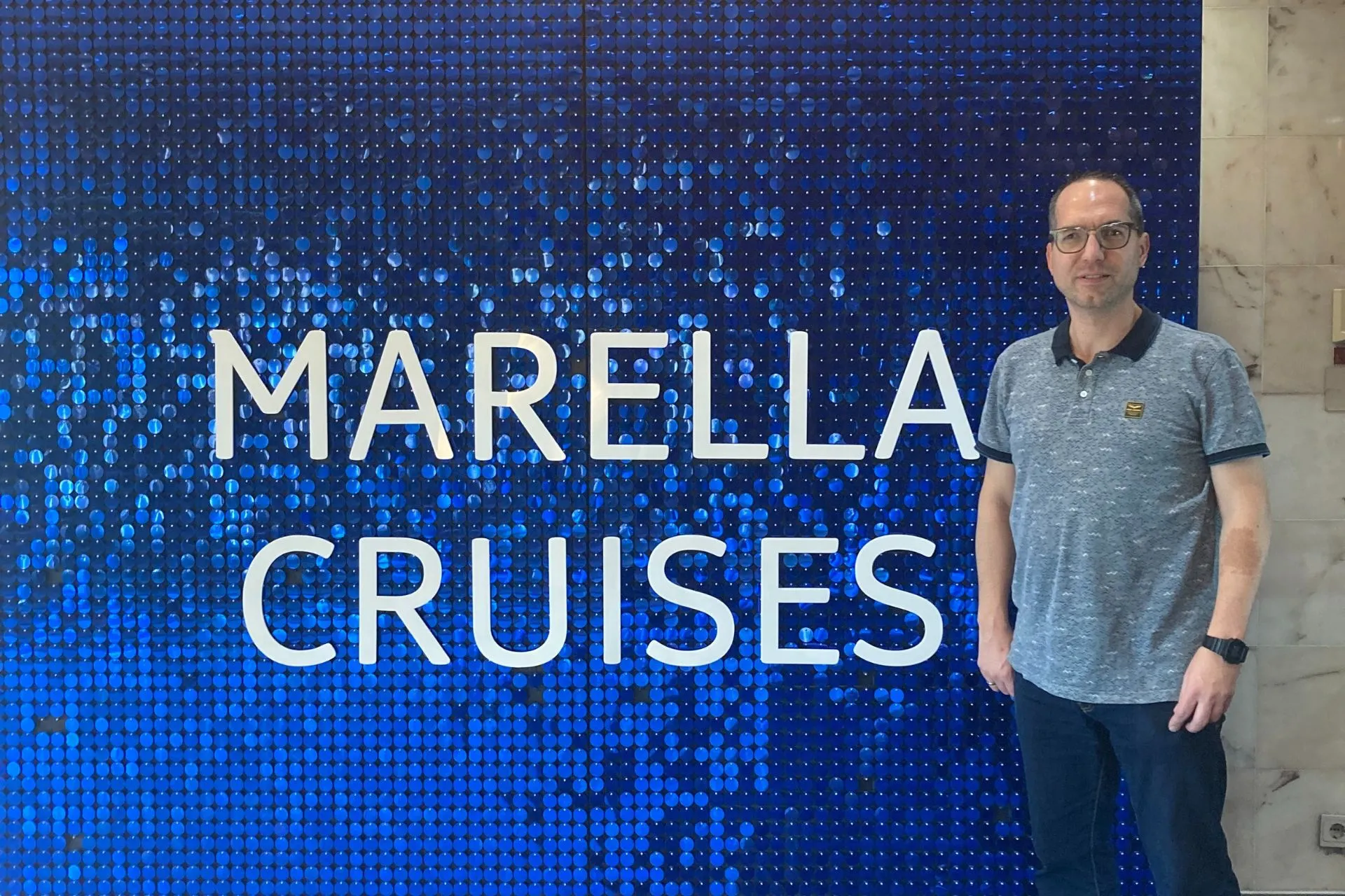 Reisadviseur Dino voor een opvallend blauw decor met de tekst 'Marella Cruises', tijdens het inschepen op de cruise.
