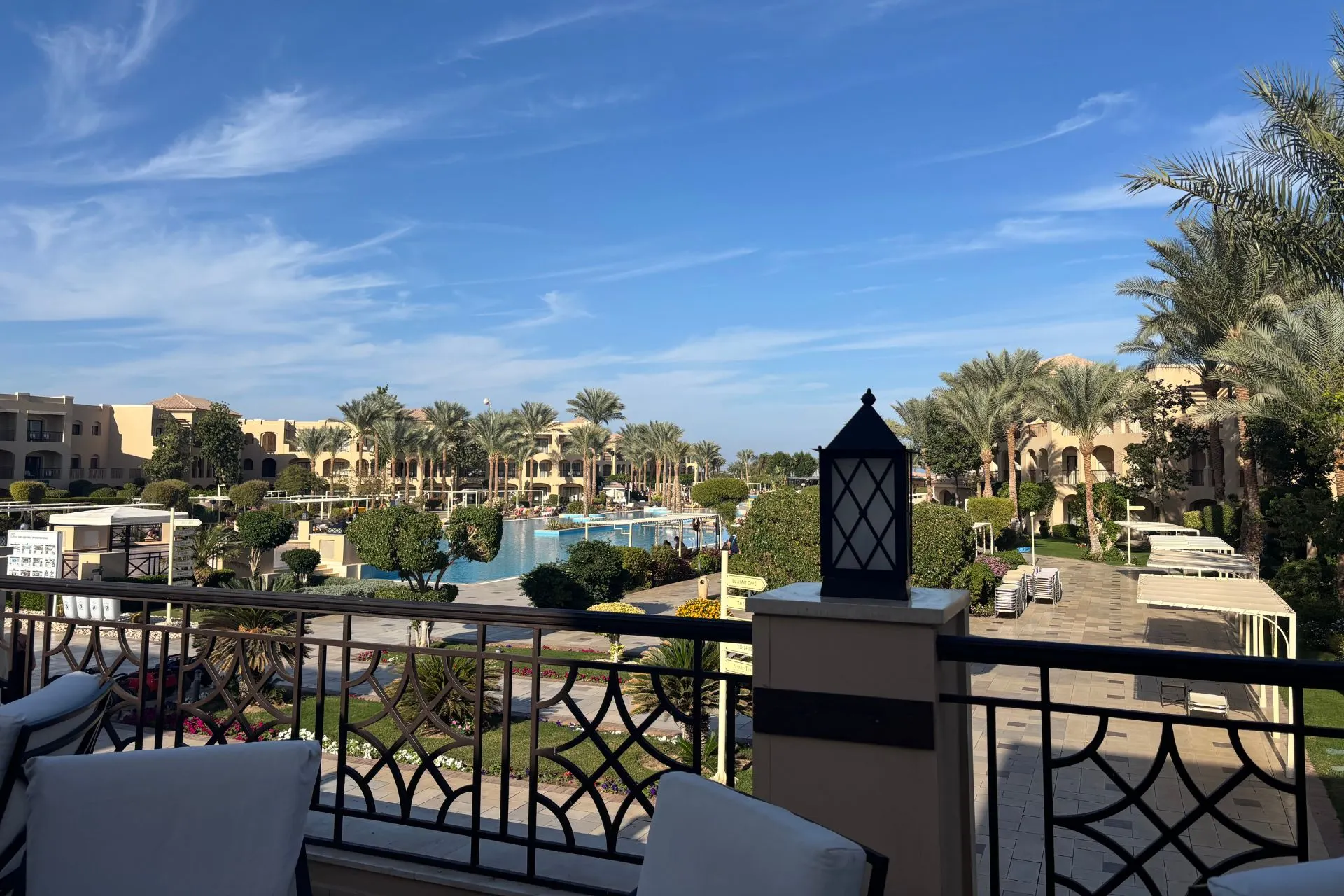 Panoramisch uitzicht vanaf een terras op een groot zwembad omringd door palmbomen en ligbedden, met elegante gebouwen en een helderblauwe lucht op de achtergrond. Op de voorgrond staat een decoratieve lantaarn op een balustrade. Dit beeld benadrukt luxe en comfort, perfect voor promotie van all-inclusive resorts en zonvakanties in Egypte.