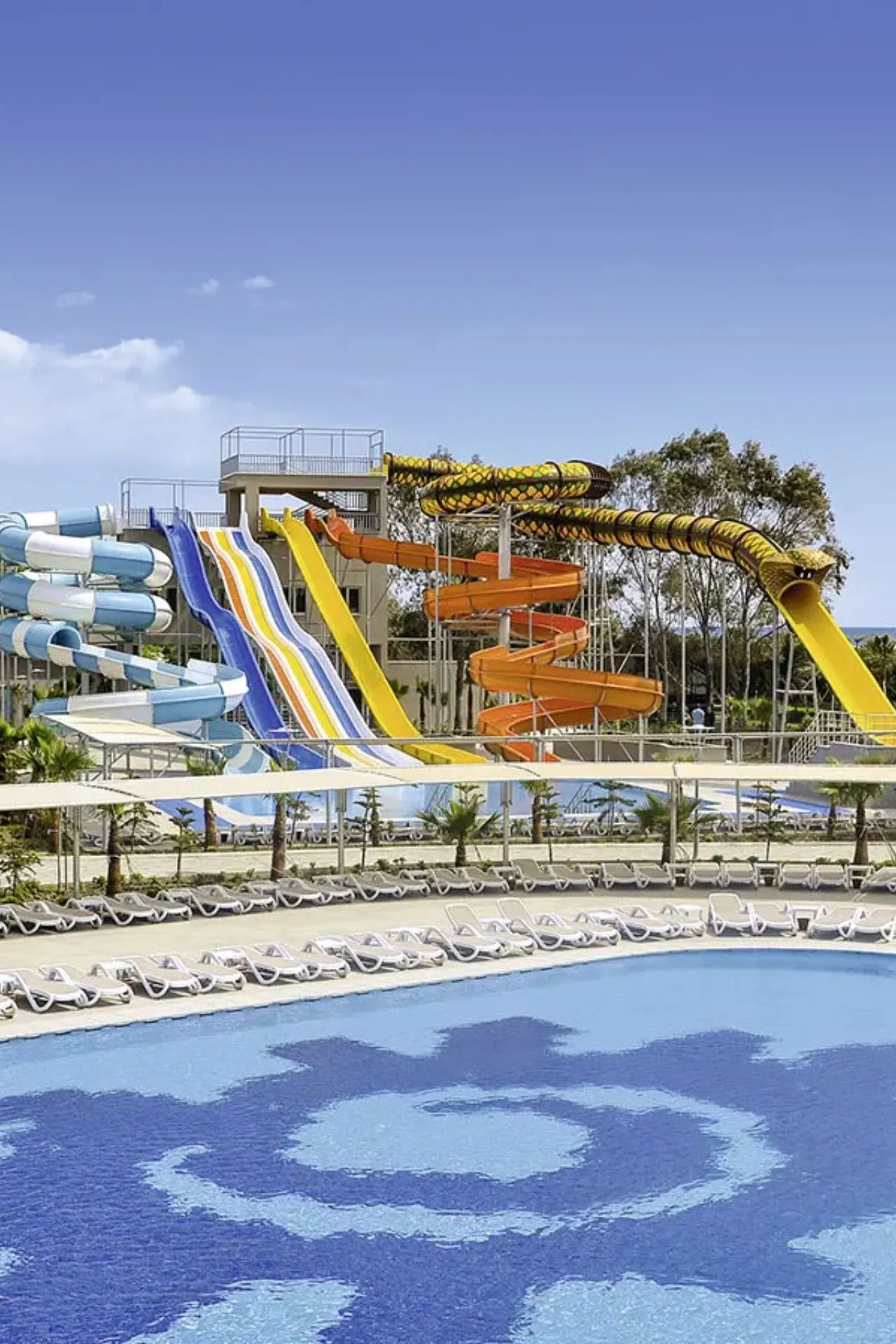 Een groot waterpark met kleurrijke glijbanen, waaronder een opvallende slangvormige glijbaan. Voor de glijbanen ligt een ruim zwembad met ligbedden rondom.