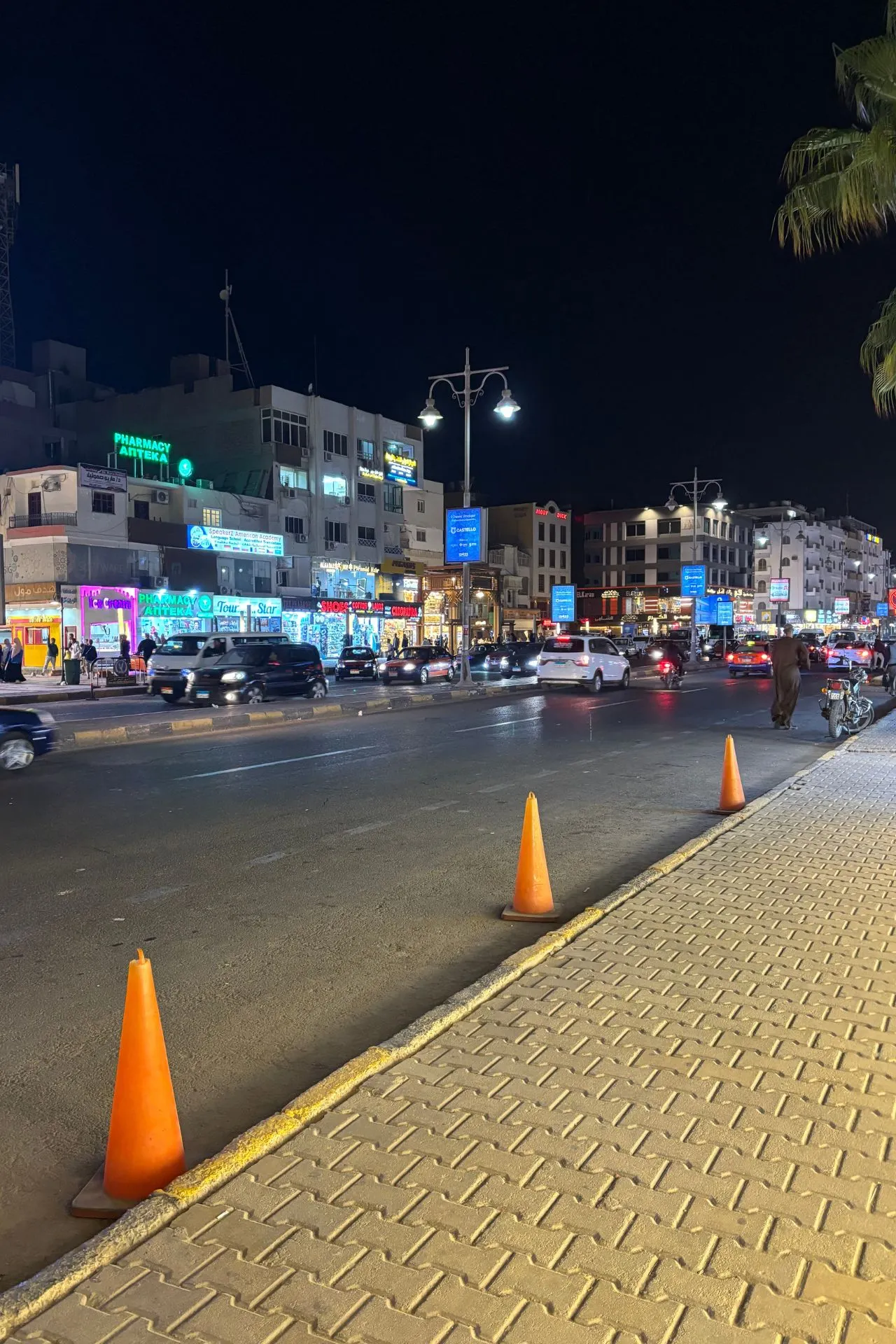 Nachtelijk straatbeeld in een levendige stad met fel verlichte winkels, neonreclames en druk verkeer op een brede weg. Auto’s rijden langs moderne gebouwen met kleurrijke lichtborden, waaronder apotheken en lokale winkels. Op de voorgrond is een betegeld trottoir zichtbaar met twee oranje verkeerskegels, terwijl straatlantaarns het gebied verlichten. Dit beeld benadrukt het bruisende nachtleven en stedelijke sfeer, ideaal voor promotie van winkelen, uitgaan en stedentrips in Egypte.