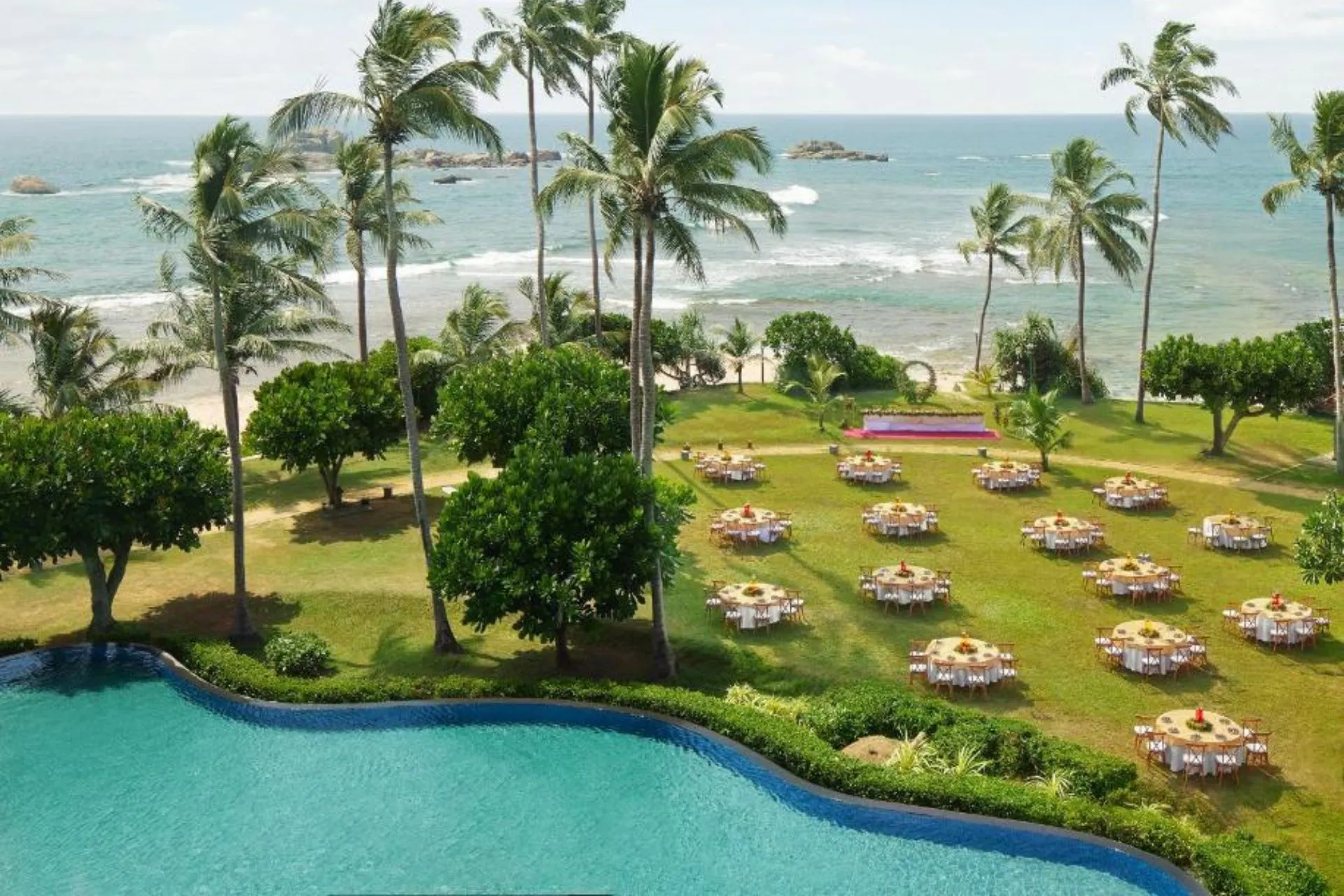  Tropische tuin van Hikka Tranz by Cinnamon met uitzicht op zee, hoge palmbomen en ronde tafels in feestelijke opstelling voor een evenement aan het strand.