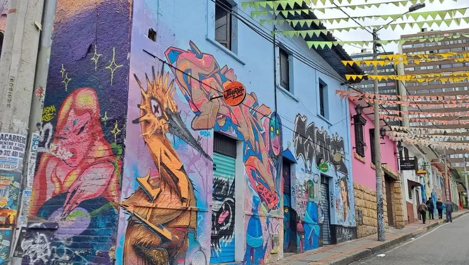 Straat met felgekleurde muurschilderingen op gebouwen, waaronder abstracte figuren en stripachtige karakters, met vlaggetjes boven de weg gespannen.