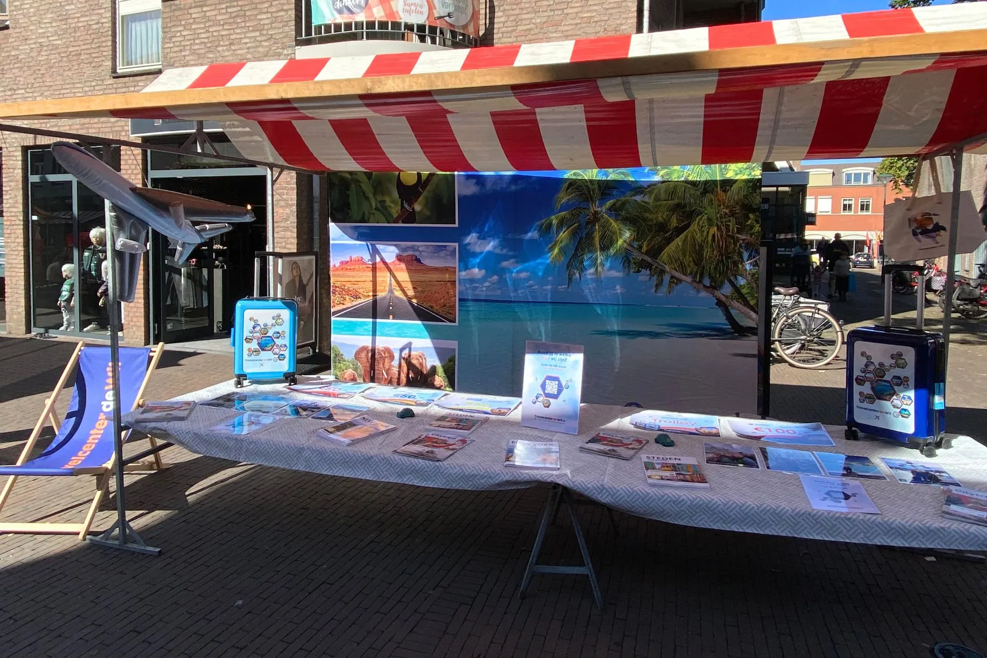 Kraampje van Travelcenter de Wit tijdens een buitenmarkt met een tropische strandachtergrond, kleurrijke foto’s van bestemmingen, een blauw ligstoeltje en promotiemateriaal op tafel zoals brochures en winactieposters.