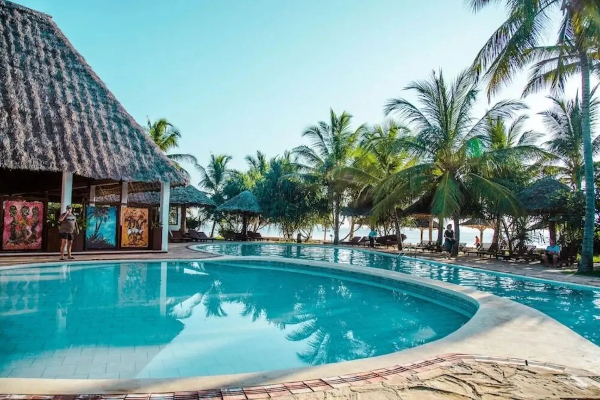 Zwembad van Jaz Adonia Beach Resort op Zanzibar, omringd door tropische palmbomen, ligbedden en traditionele gebouwen met rieten daken. Dit familievriendelijke resort aan Uroa Beach biedt een ontspannen vakantiesfeer, moderne faciliteiten en een prachtige ligging vlak bij het hagelwitte strand en de Indische Oceaan.