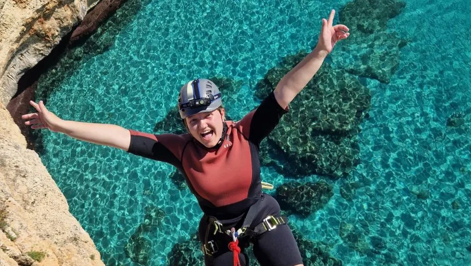 Vrouw in wetsuit en klimuitrusting hangt lachend met gespreide armen aan een rood touw boven helder turquoise zeewater met zichtbare rotsen eronder.