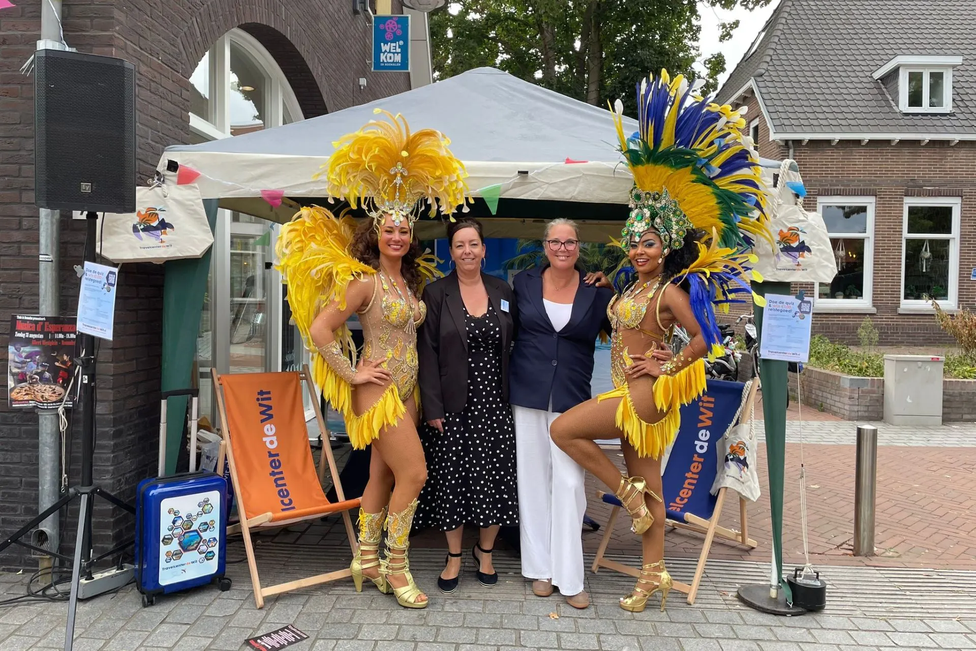 Twee medewerkers van Travelcenter de Wit poseren bij de kraam samen met twee sambadanseressen in opvallende geel-blauwe kostuums tijdens Thuis in Rosmalen 2025.