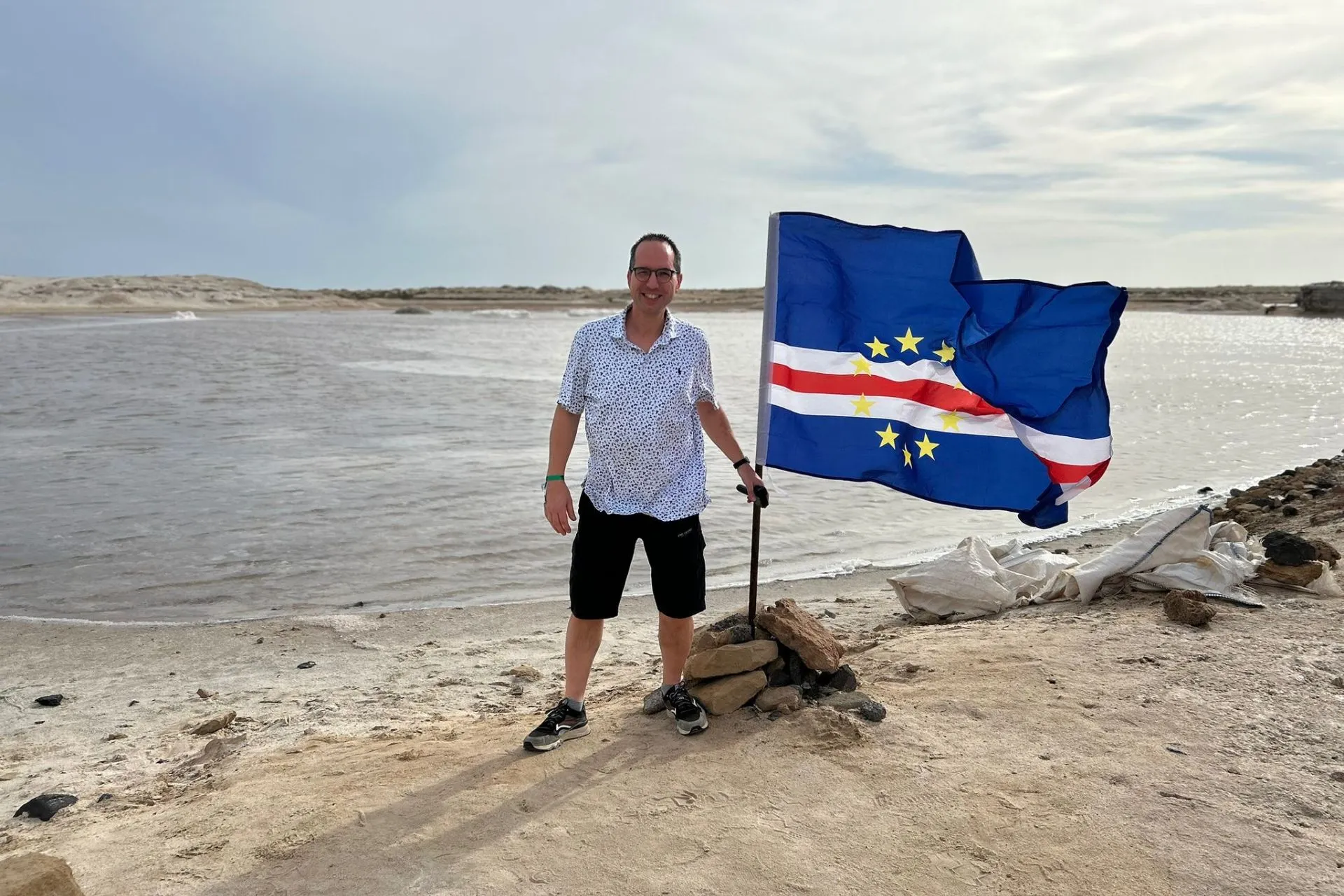 Persoon aan de rand van een lagune op Kaapverdië met een Kaapverdische vlag die stevig wappert in de wind, omgeven door een droog en ruig landschap. De combinatie van water, zand en open ruimte weerspiegelt het unieke karakter van een vakantie Kaapverdië, waar natuur en cultuur samenkomen. Dit beeld past goed bij een actieve reis Kaapverdië, waarbij je naast stranden ook bijzondere natuurgebieden, zoutmeren en afgelegen plekken ontdekt die typerend zijn voor eilanden zoals Sal en Boa Vista.