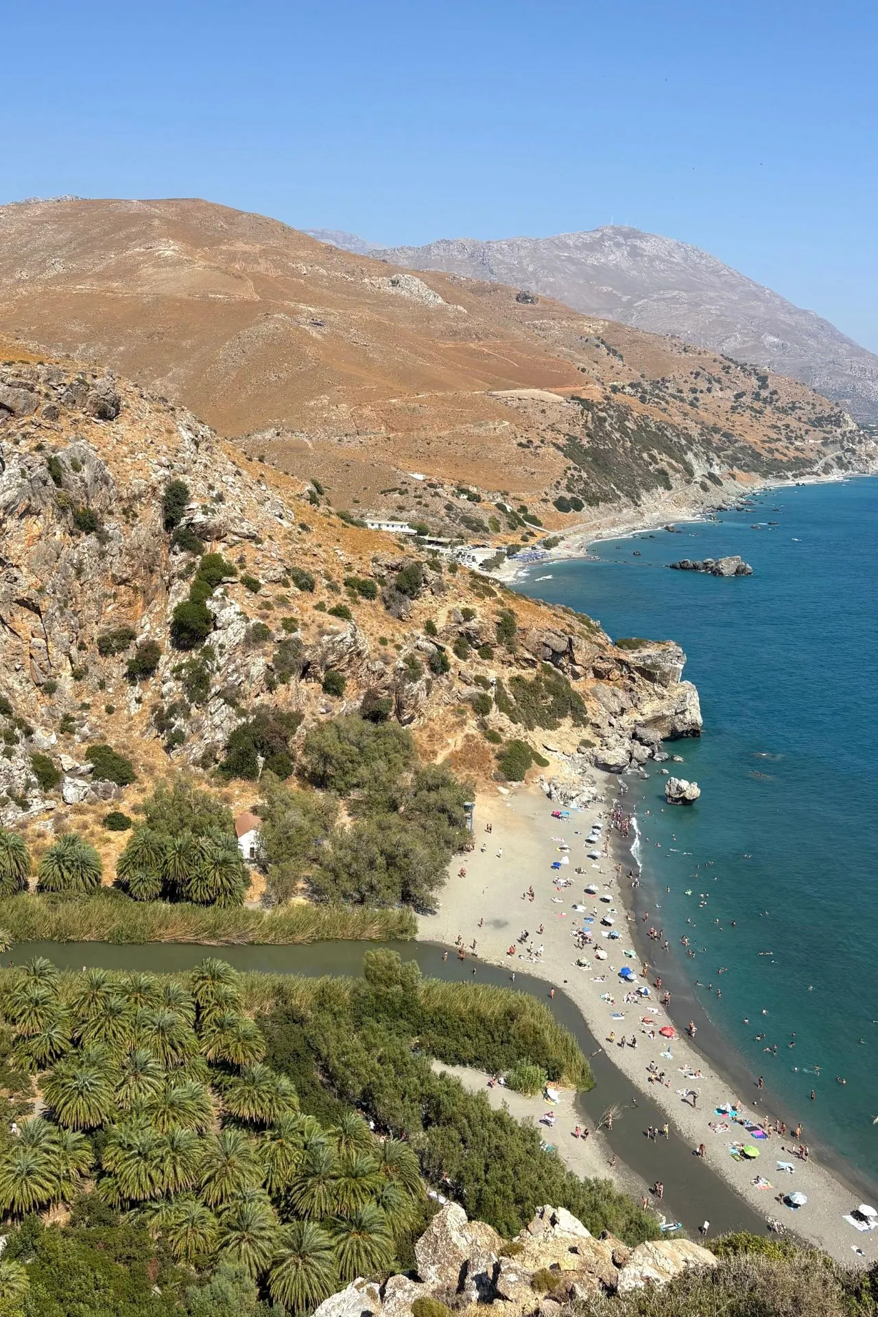 Panoramisch uitzicht over Preveli Beach met turquoise zee, palmbomen en een rivier die uitmondt in het strand, omringd door droge heuvels.